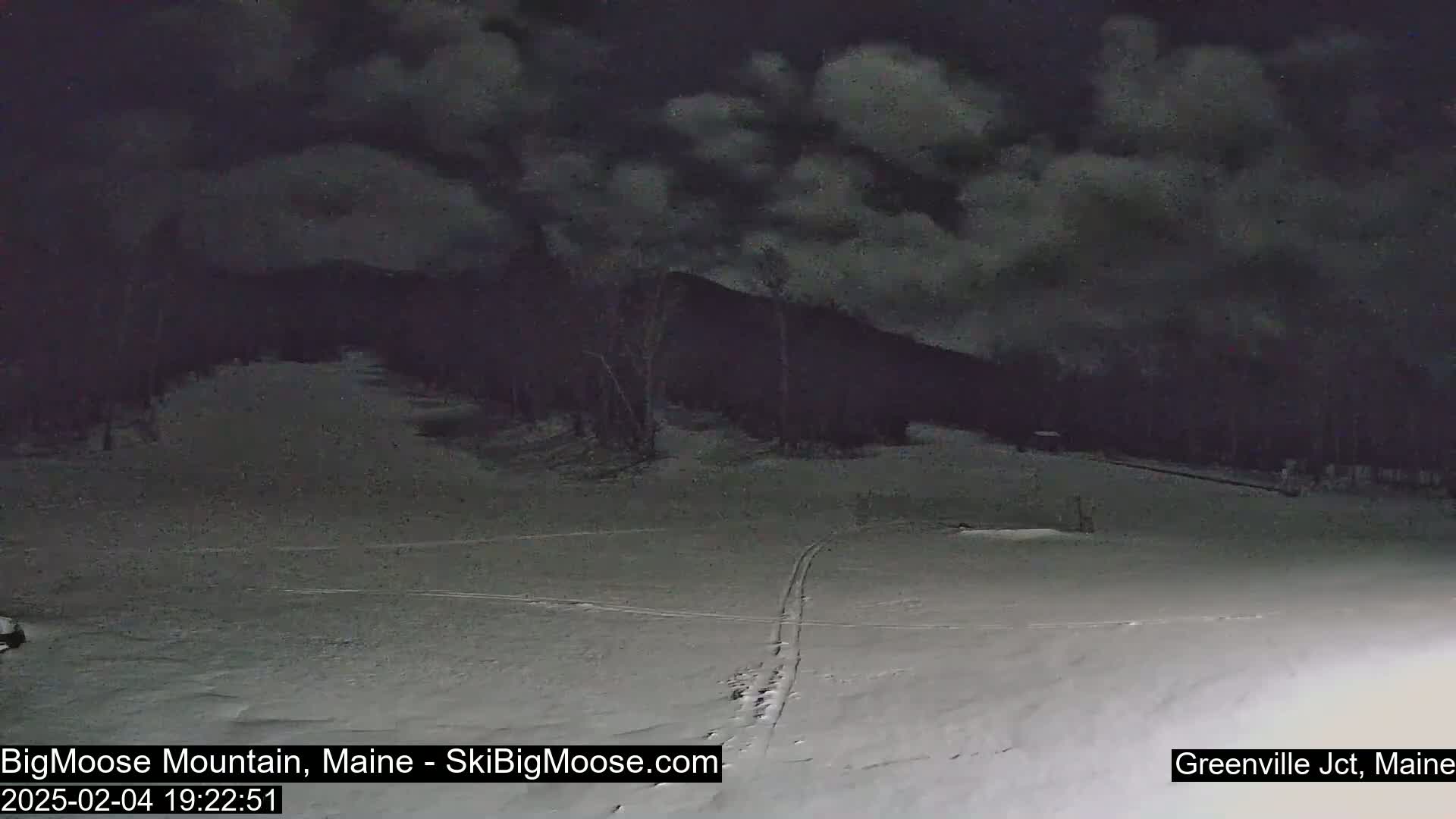 A snow-covered landscape at night under a cloudy sky shows a dark mountain in the background and faint ski tracks on the foreground.
