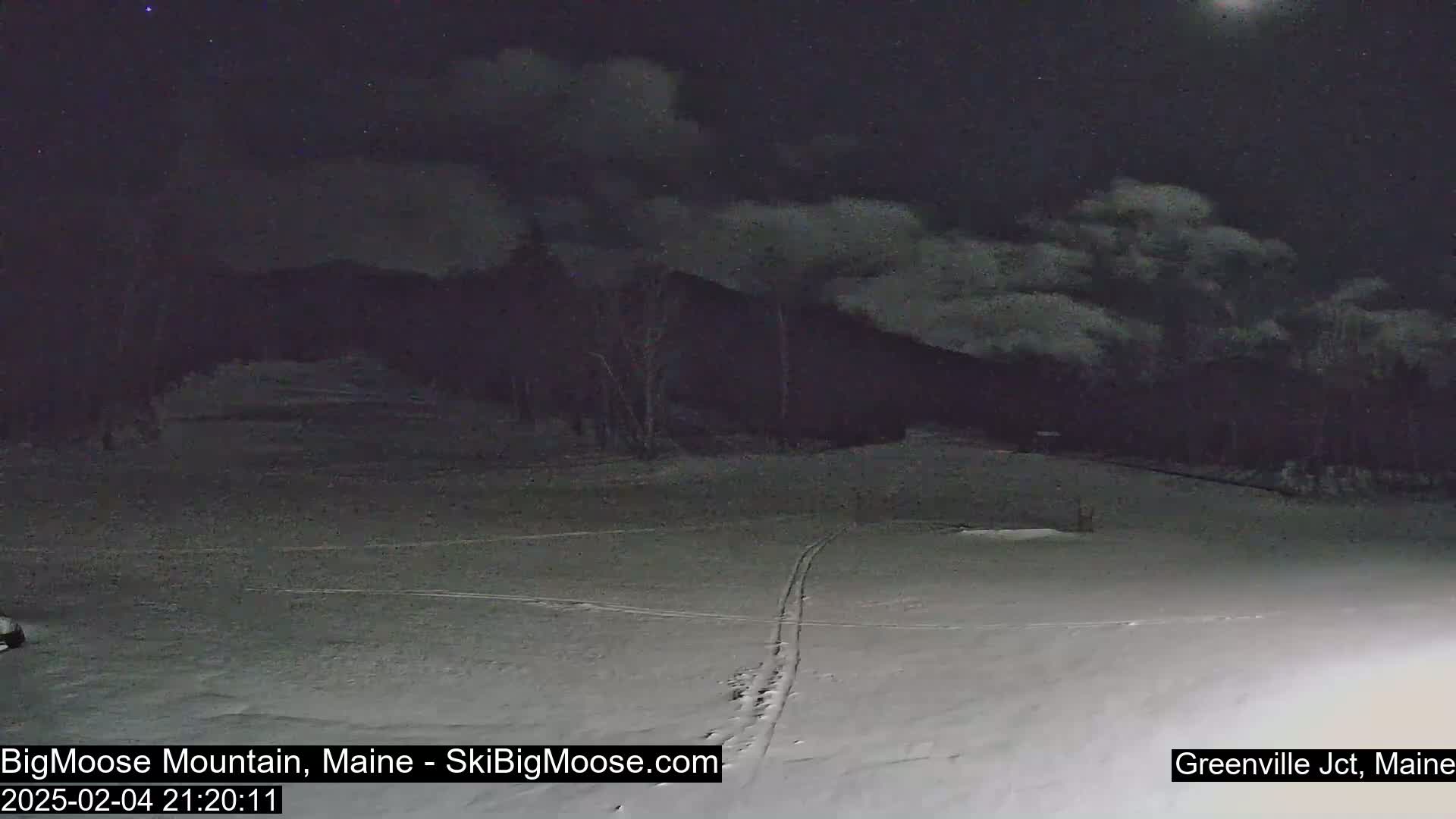 A snow-covered area at night with a dark, cloudy sky and trees in the background, showing faint ski tracks on the snow.