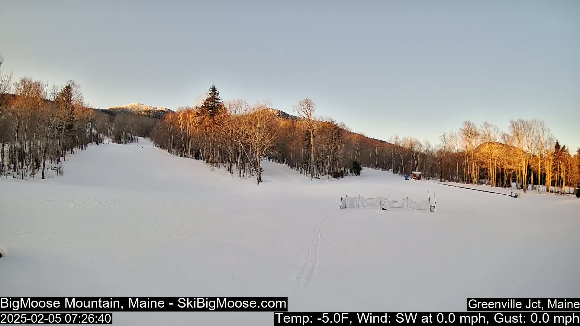 A snow-covered ski slope at sunset, with bare trees and mountains in the background under a clear sky.
