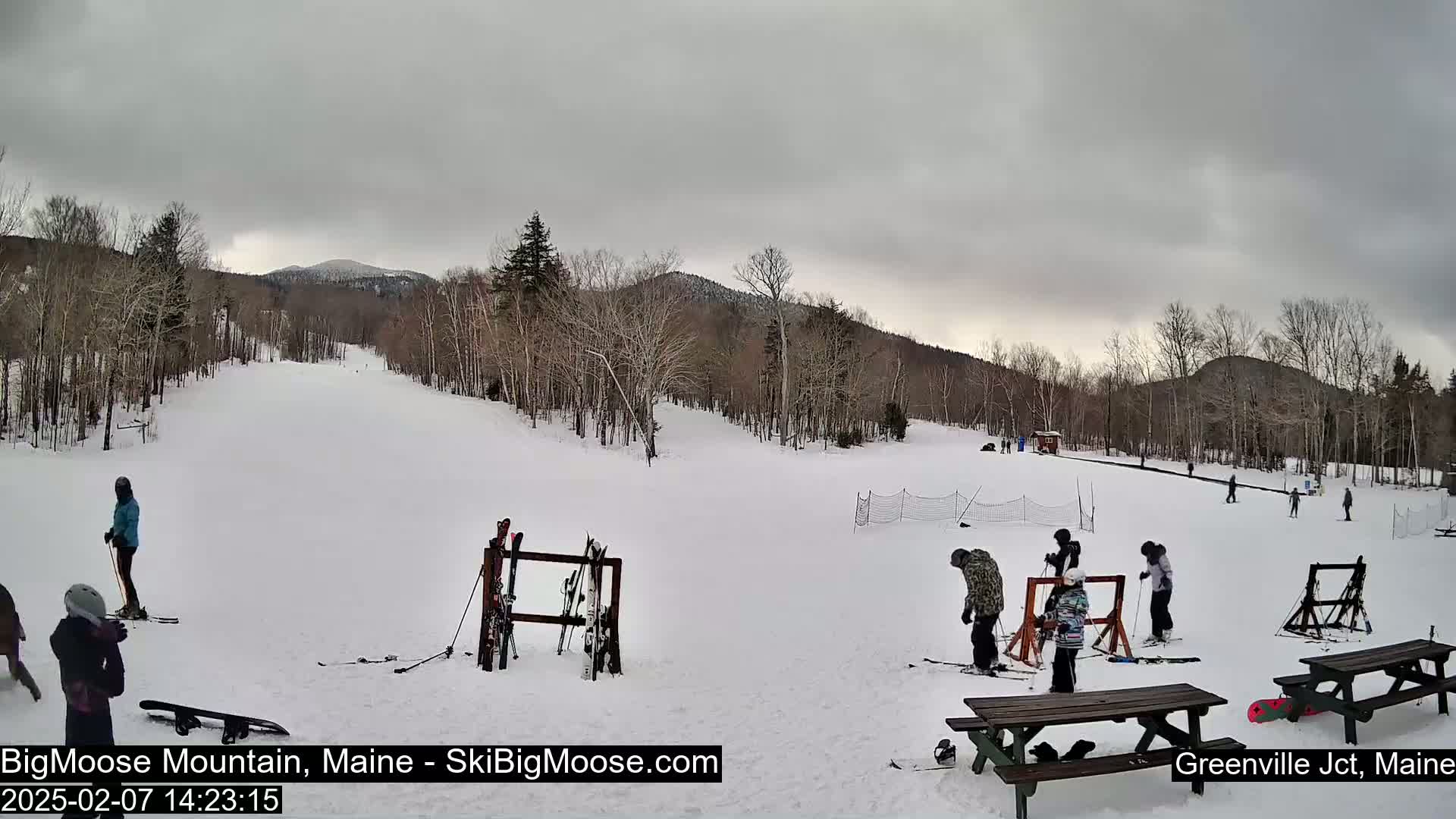 A snowy ski slope with several skiers and snowboarders, ski racks, picnic tables, and trees under an overcast sky.