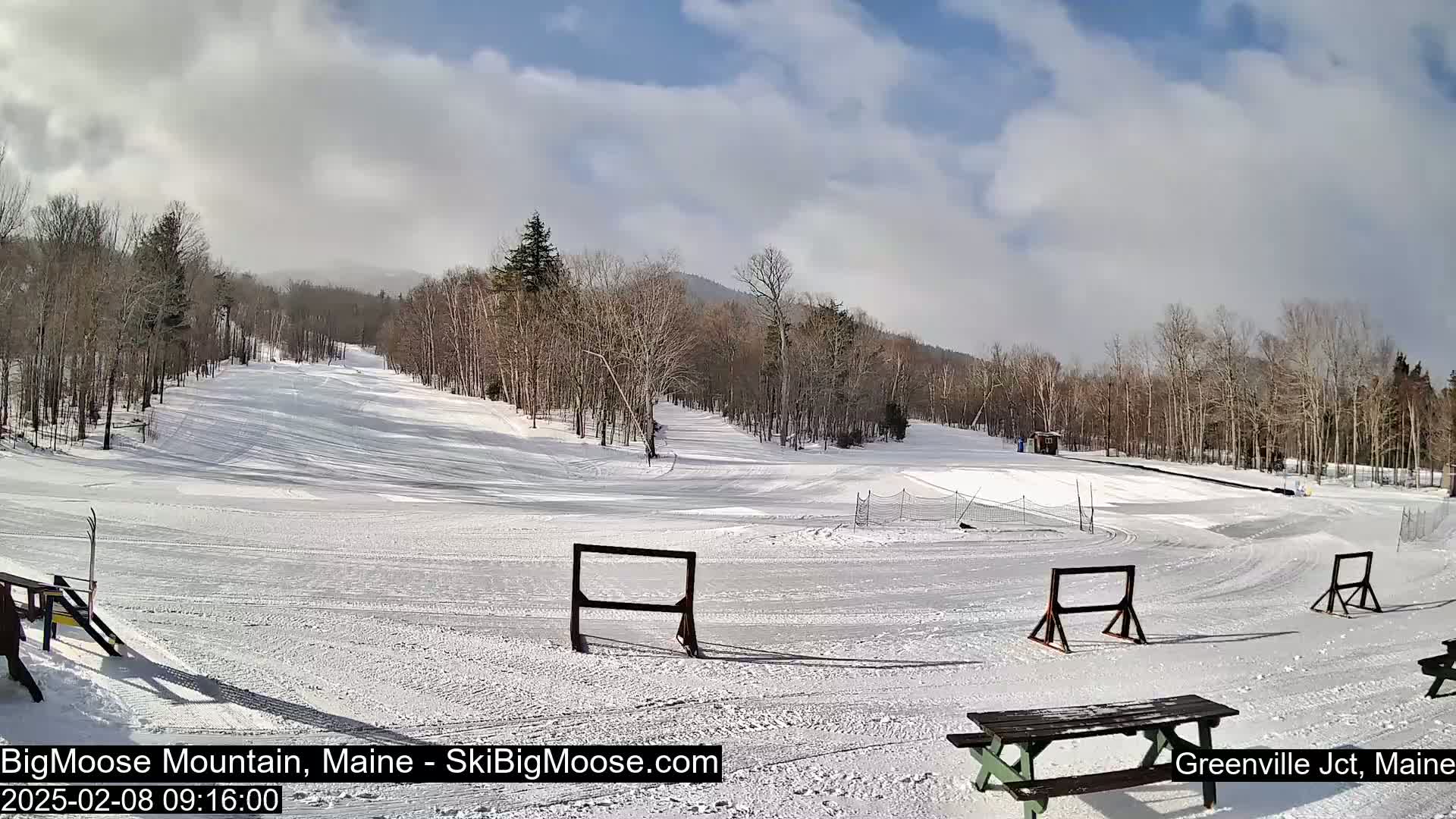 A snow-covered ski slope with several wooden barriers and a picnic table is visible under a partly cloudy sky.