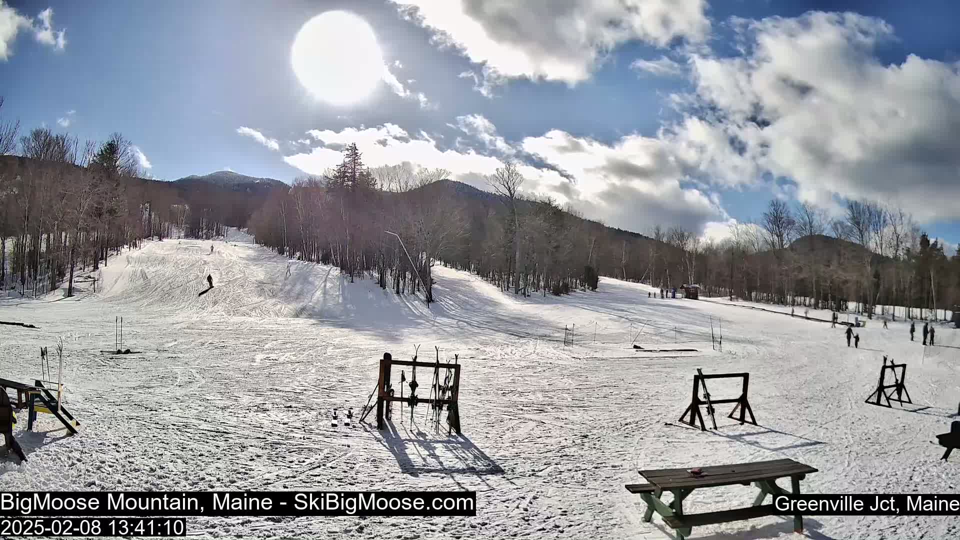 A sunny day at a snow-covered ski resort with several people skiing and snowboarding down a slope, surrounded by leafless trees and distant mountains.