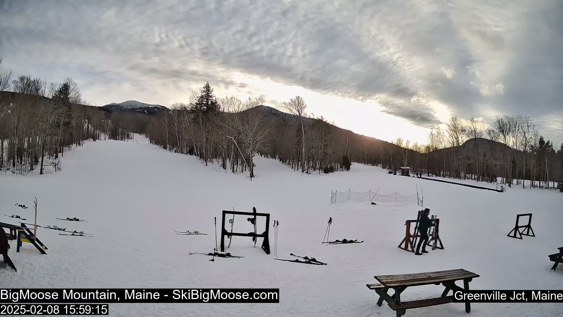 A snow-covered ski slope with several pairs of skis leaning against racks, a person near a rack, and a picnic table in the foreground, all set against a backdrop of bare trees and mountains under a cloudy sky.