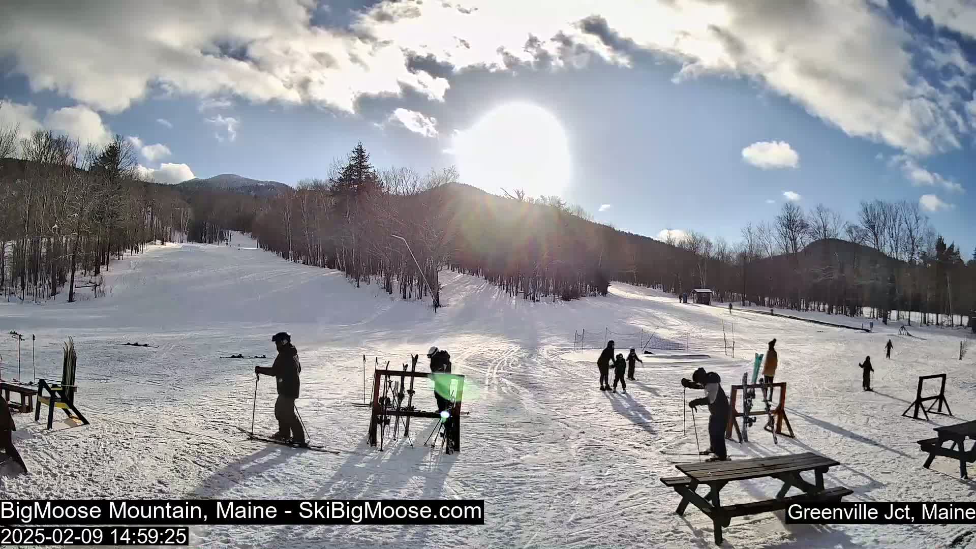 A sunny, partly cloudy day at a snow-covered ski slope with several skiers and snowboarders present,  with trees and mountains in the background.