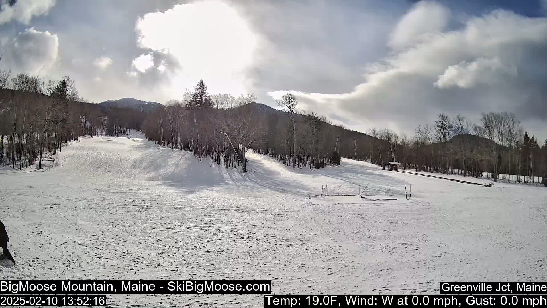A snow-covered ski slope with sparsely wooded hills in the background under a partly sunny sky.