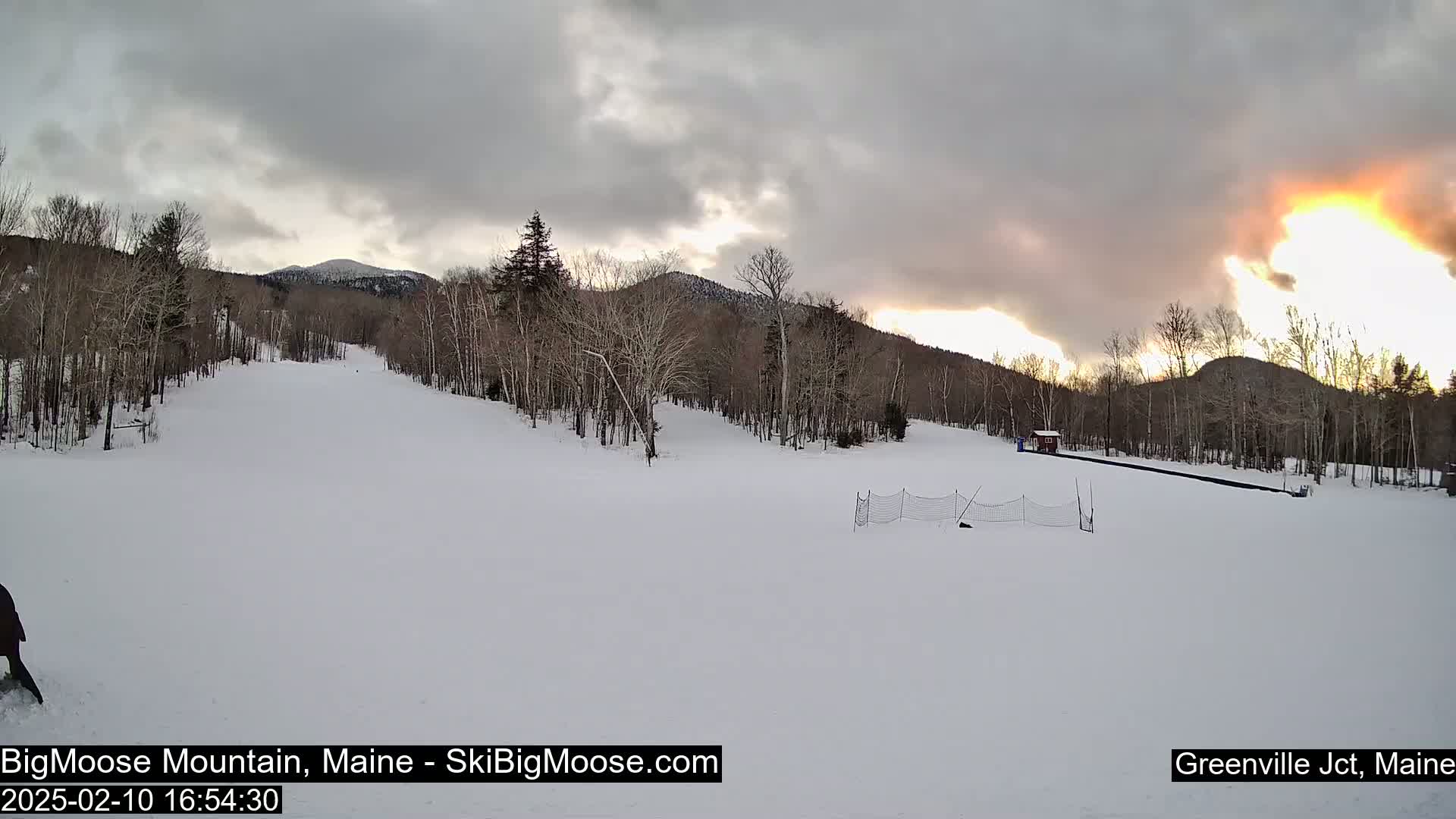 A snow-covered ski slope is nestled among bare, snow-dusted trees under a cloudy sky with a bright sunset breaking through the clouds in the distance.