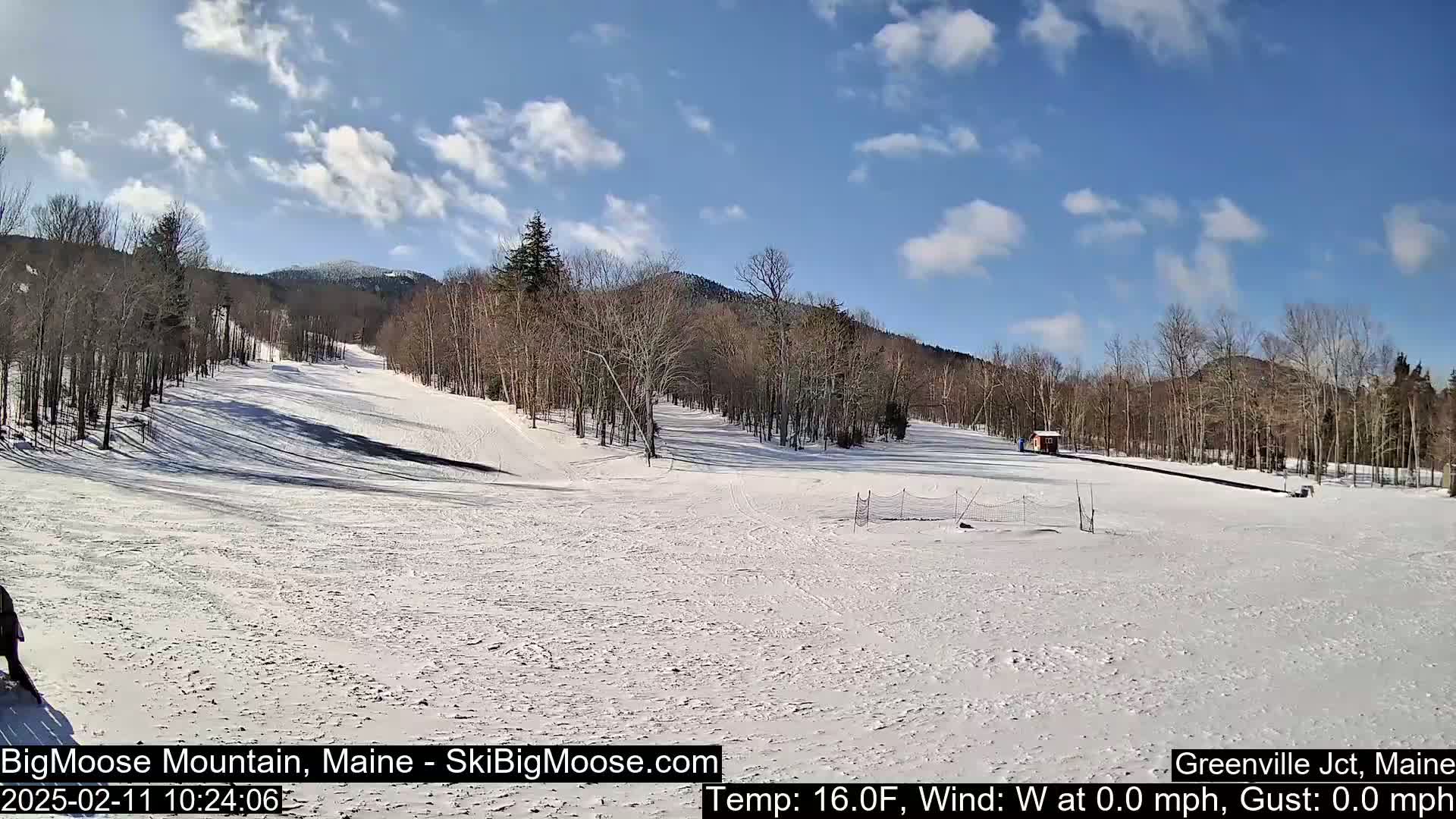 A snow-covered ski slope stretches towards snow-capped mountains under a mostly sunny sky.