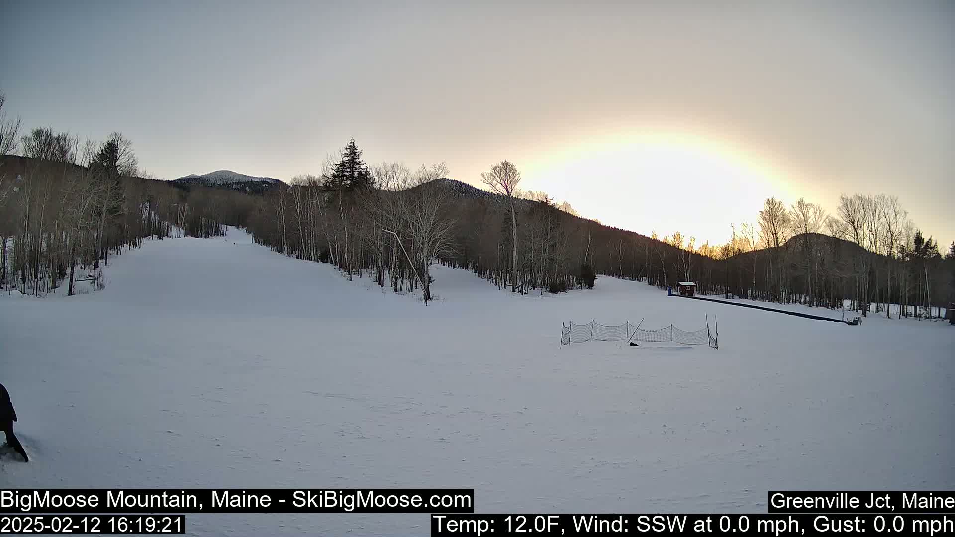 A snow-covered ski slope at sunset, with bare trees lining the hills in the background under a clear sky.
