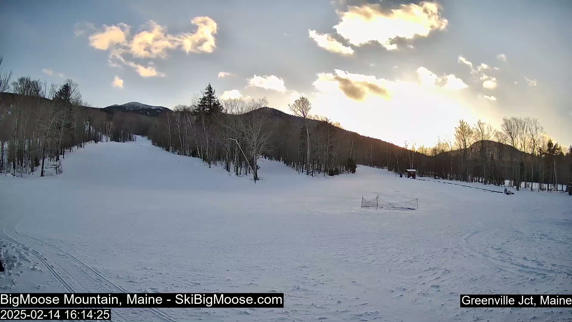 A snow-covered ski slope is nestled amongst bare trees under a partly cloudy sunset sky.