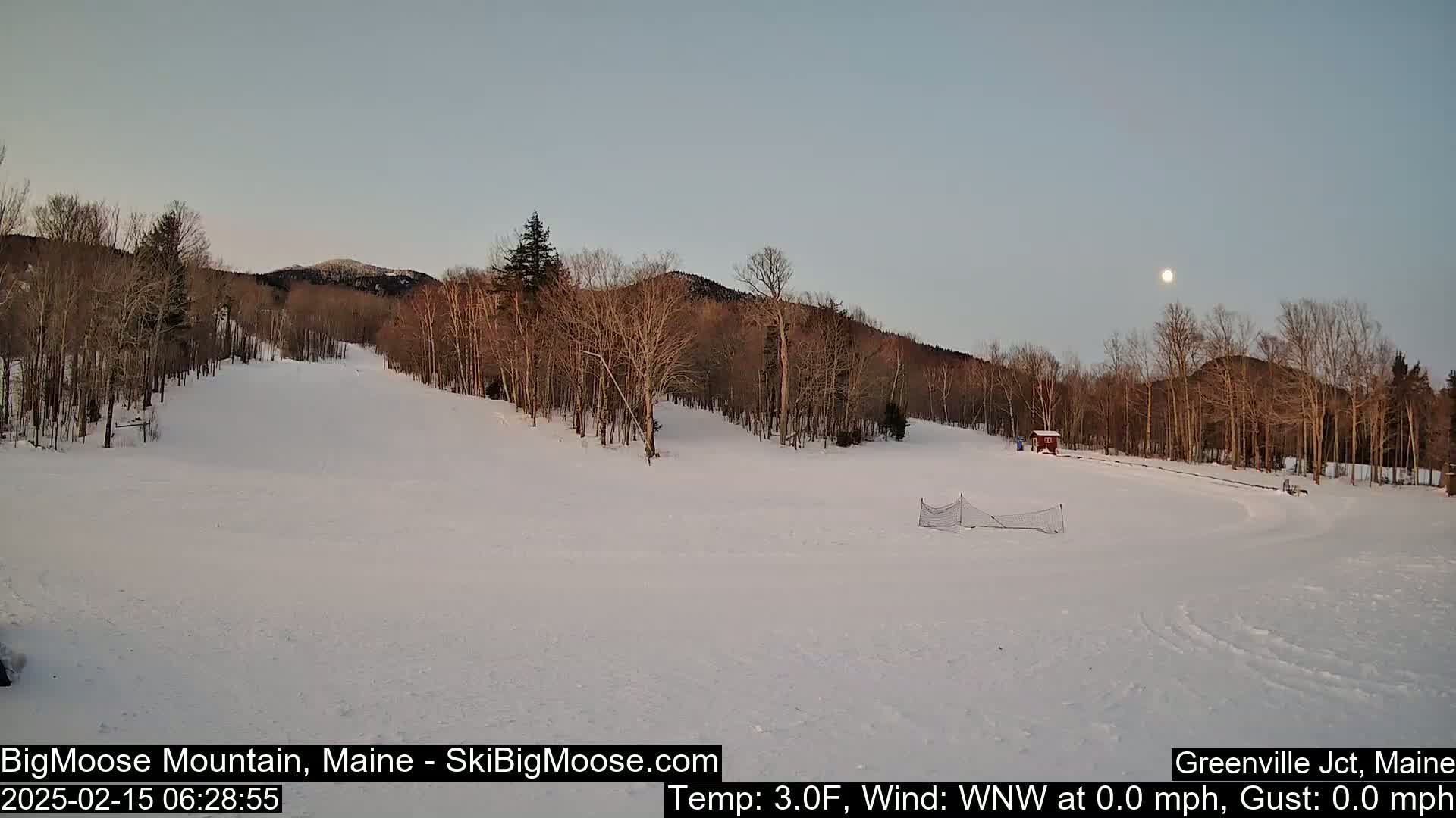 A snow-covered ski slope is visible under a twilight sky with a full moon, surrounded by bare deciduous trees and a mountain in the background.