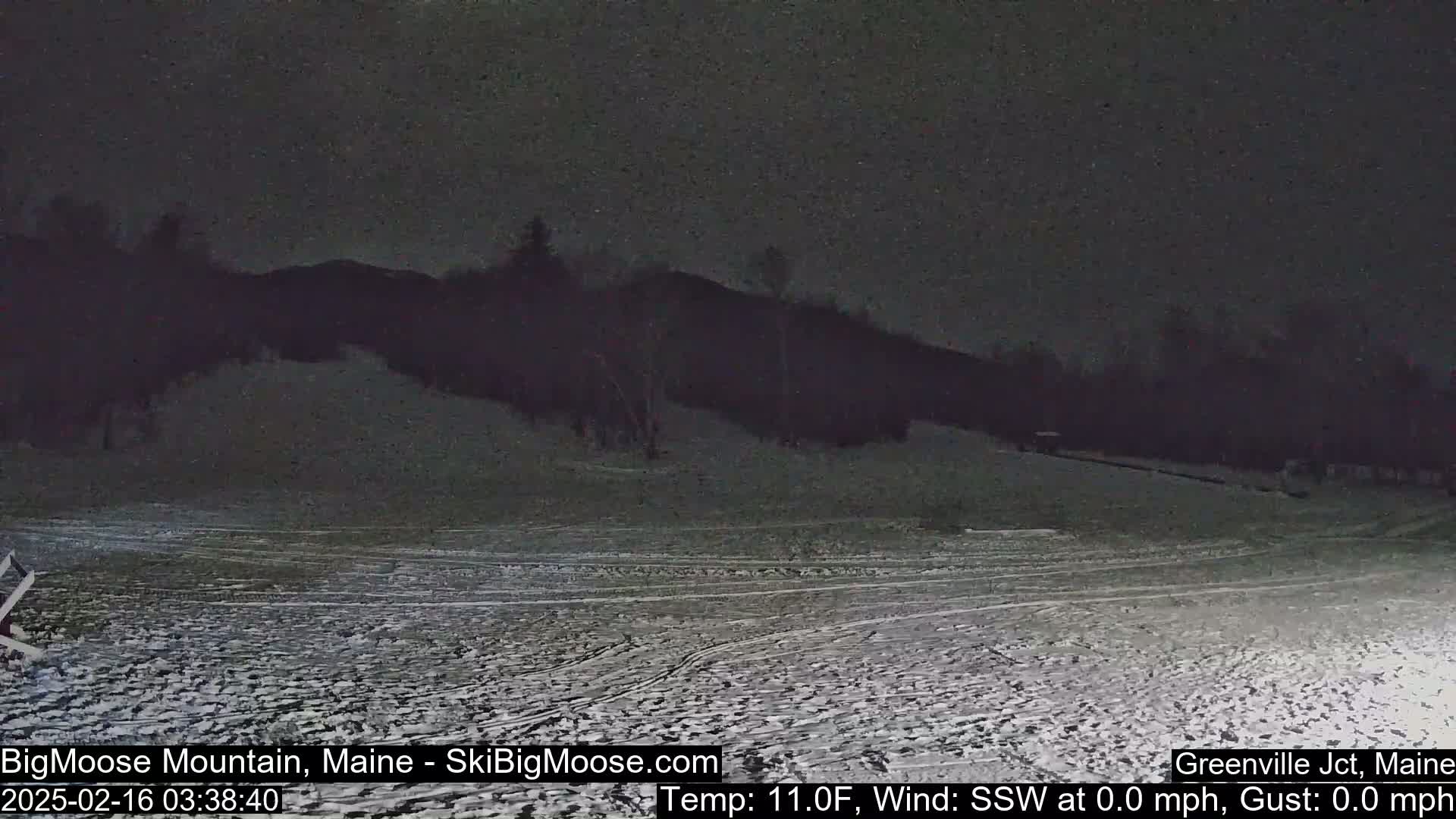 A snow-covered, dark landscape at night shows a hill silhouetted against a cloudy sky.