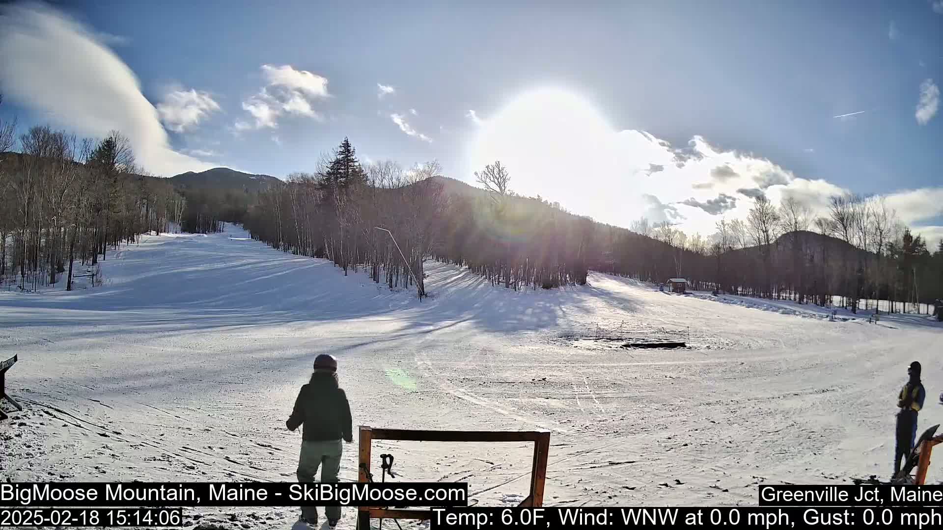 A sunny day at a snow-covered ski slope, with a few people present and trees and mountains in the background.