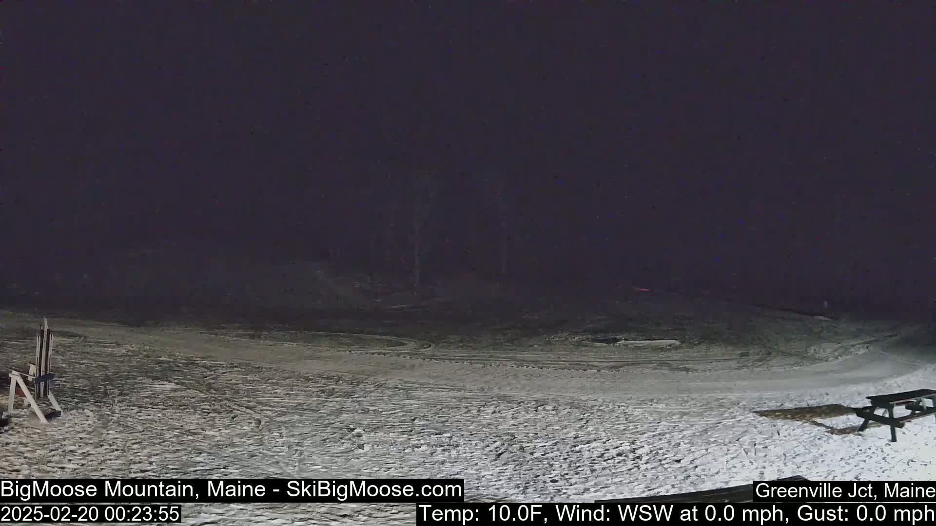 A snow-covered, nighttime scene shows a mostly empty ski slope with a picnic table and a small structure in the lower left.