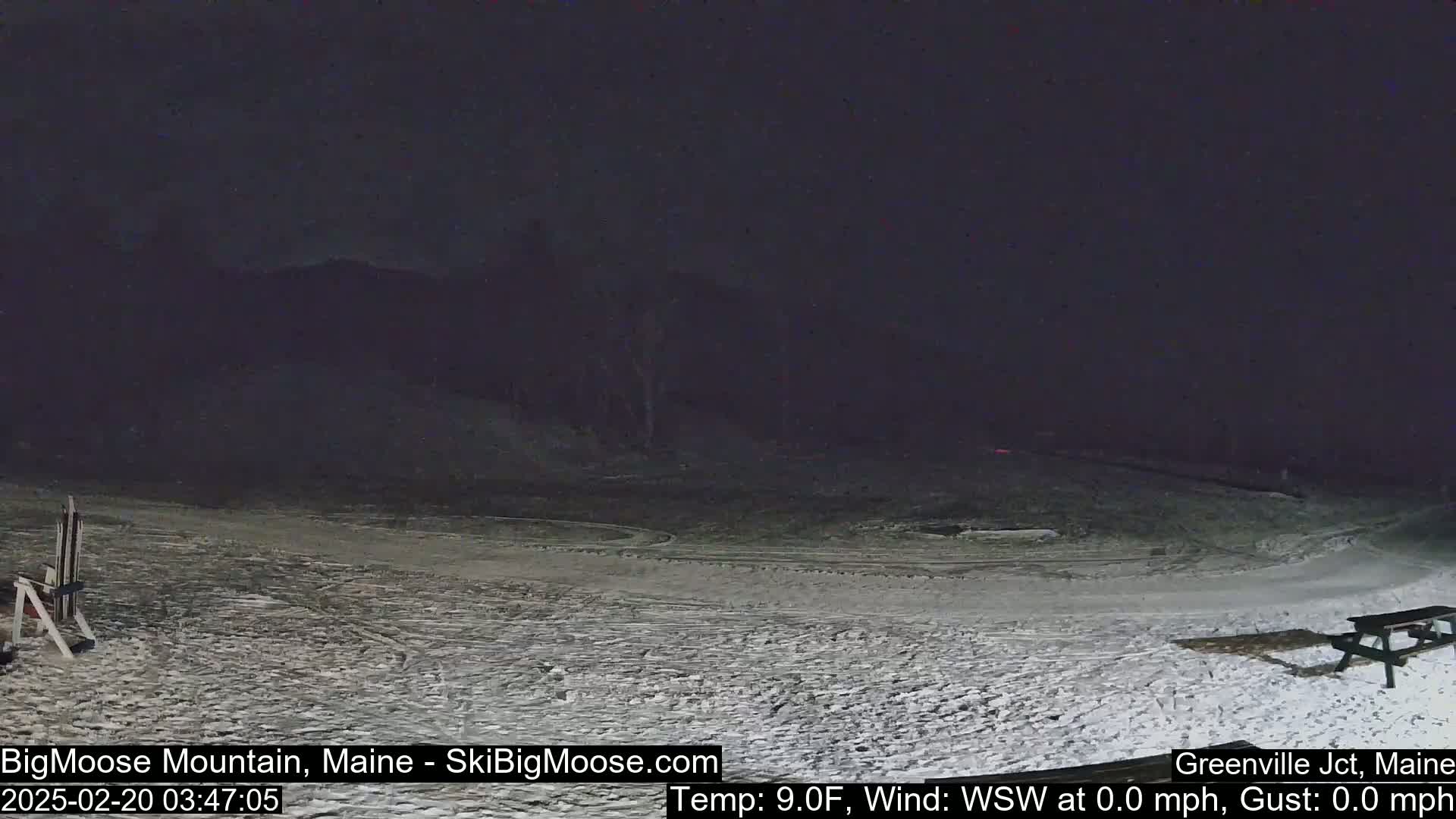 A snow-covered nighttime scene shows a mostly empty ski slope with a picnic table and ski equipment in the foreground, under a dark, clear sky.