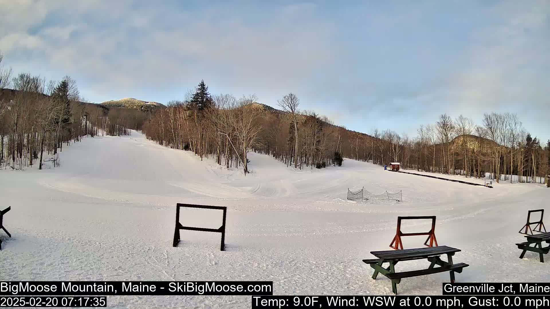A snow-covered ski slope with several picnic tables and bare trees under a partly cloudy sky.