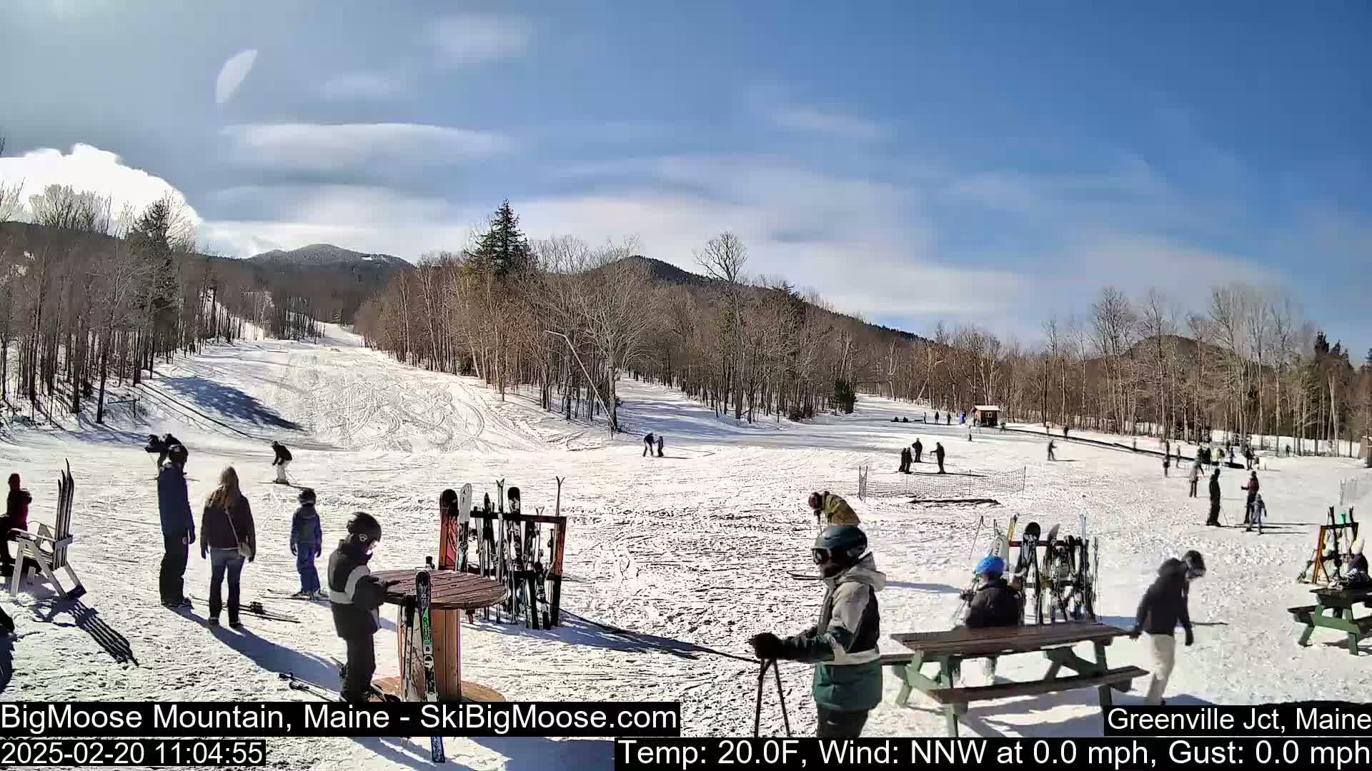 A sunny, snow-covered ski slope with several people skiing and standing around, some near picnic tables and ski racks, with bare trees and mountains in the background.