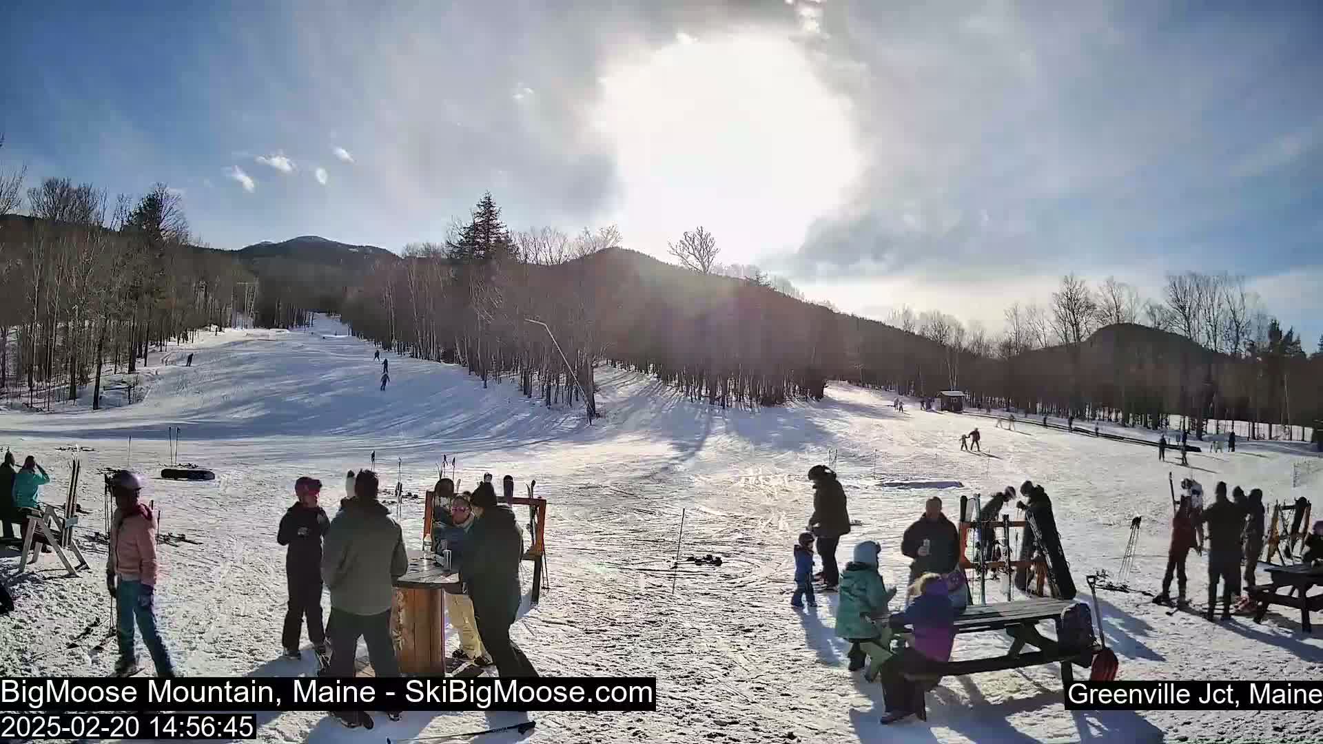 A sunny day at a snow-covered ski resort shows skiers and snowboarders on a slope, with people gathered near picnic tables at the base of the hill.