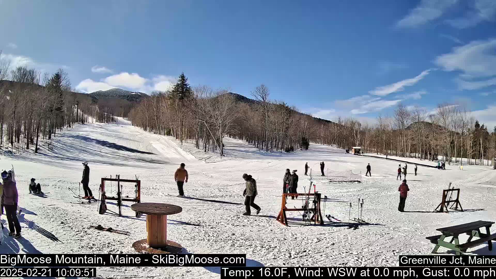 A sunny, snow-covered ski slope with several people skiing and standing around.