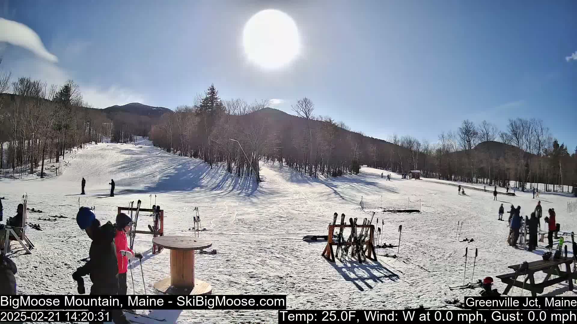 A sunny, snow-covered ski slope with several skiers and snowboarders, ski racks, and people gathered near a picnic table.