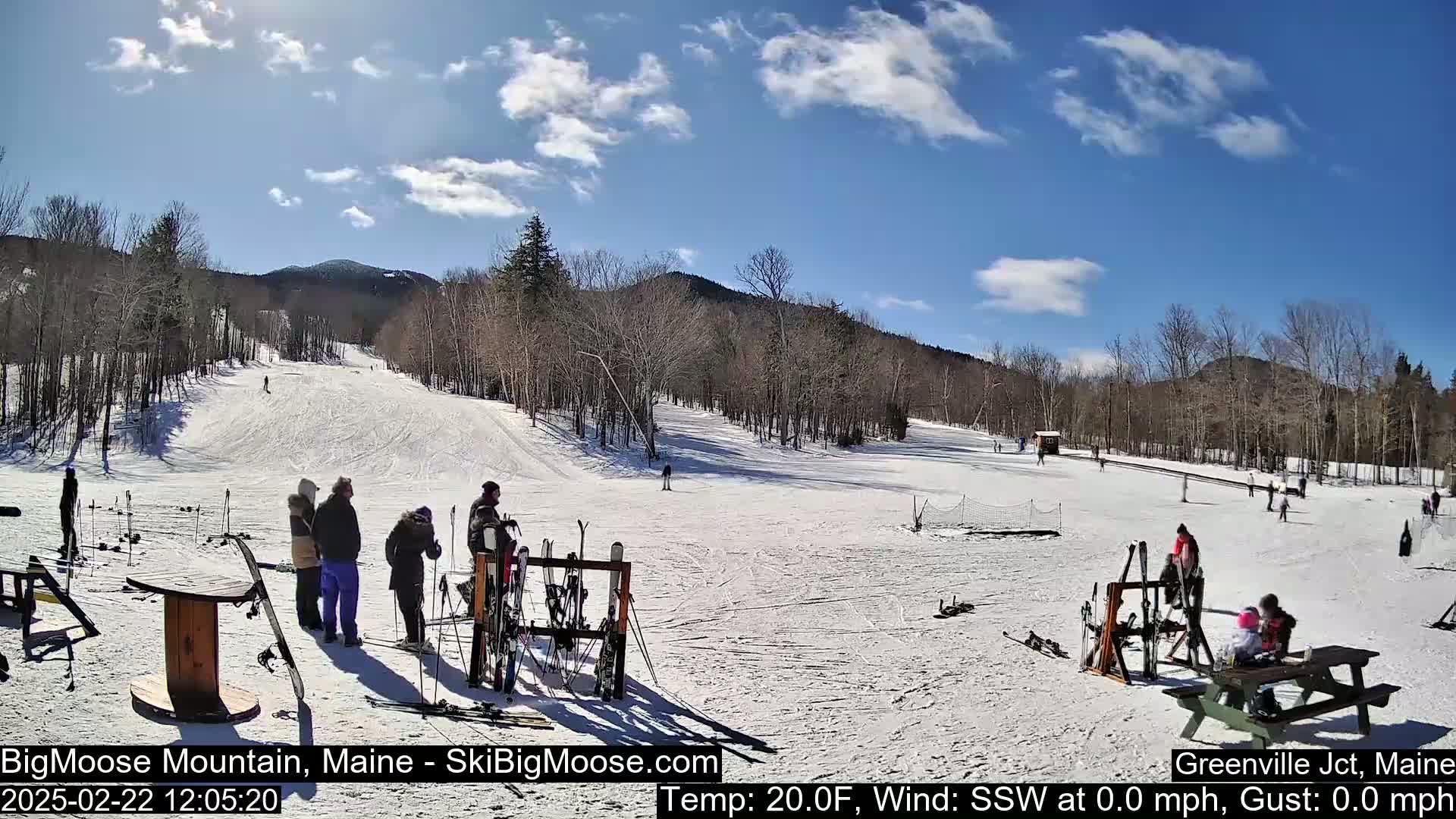 A sunny, snow-covered ski slope with several people standing near ski racks and a picnic table, surrounded by bare trees and mountains in the background.