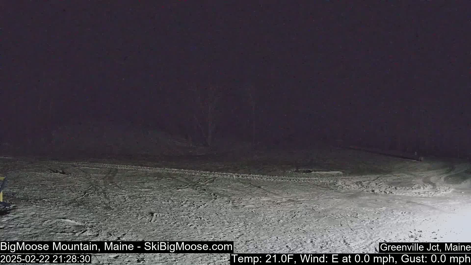 A snow-covered, dark area at night with tire tracks visible on the ground and a calm wind.