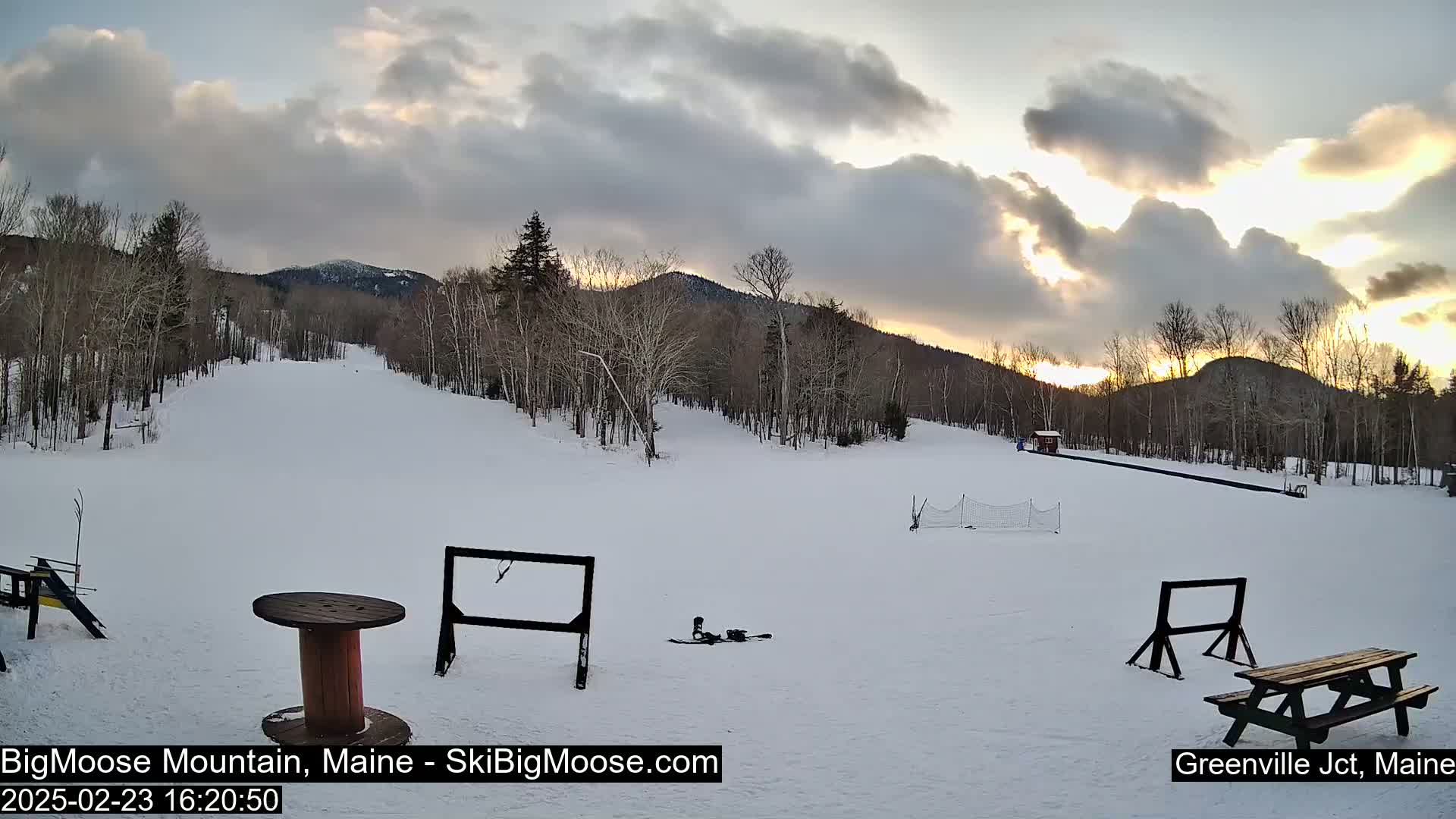 A snowy ski slope at sunset, under a partly cloudy sky, is surrounded by bare deciduous trees and distant mountains.