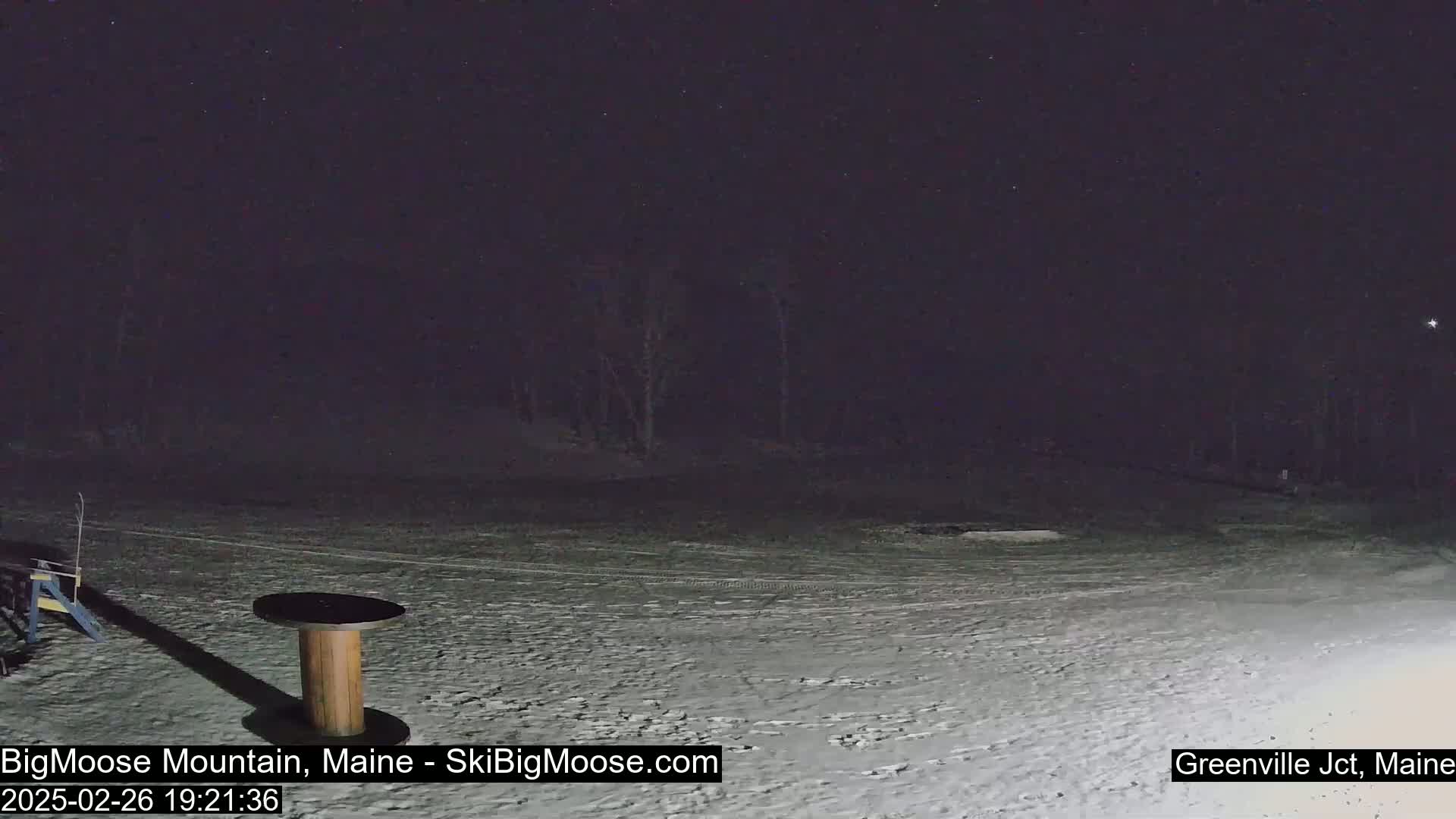 A snow-covered, dark area at night with a wooden spool-like table and some dark trees in the background under a clear, starry sky.