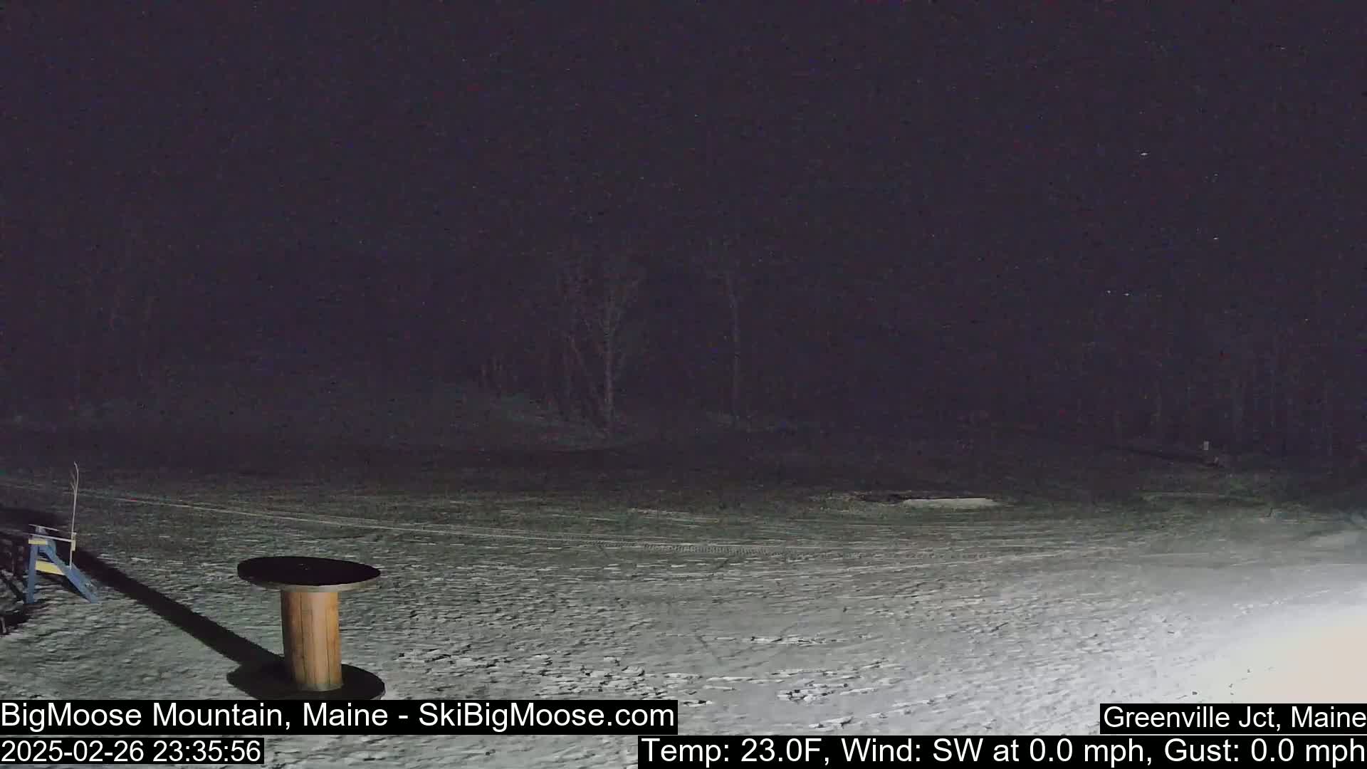 A snow-covered, dark nighttime scene shows a wooden spool table and a small amount of equipment in the foreground, with a dark treeline in the background under a clear, dark sky.