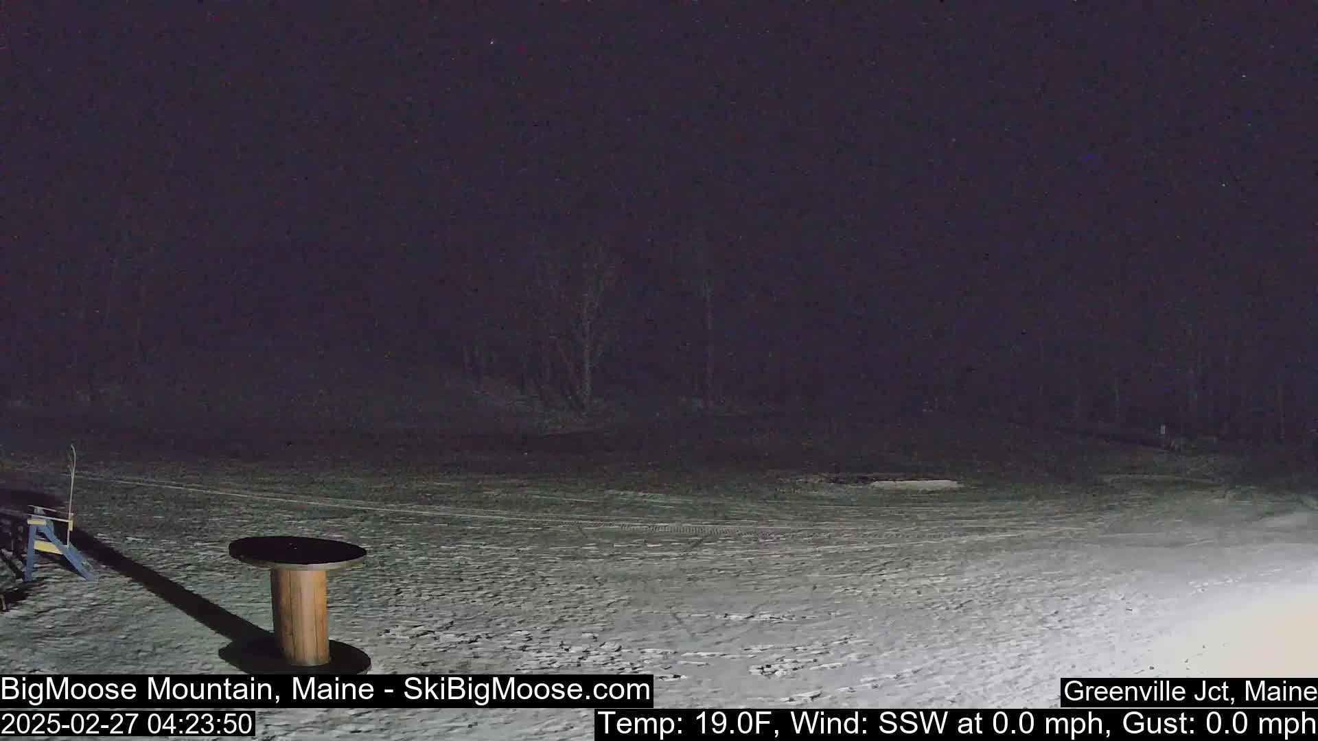 A snow-covered, dark nighttime scene shows a wooden spool table and a small portion of a structure under a mostly clear sky.