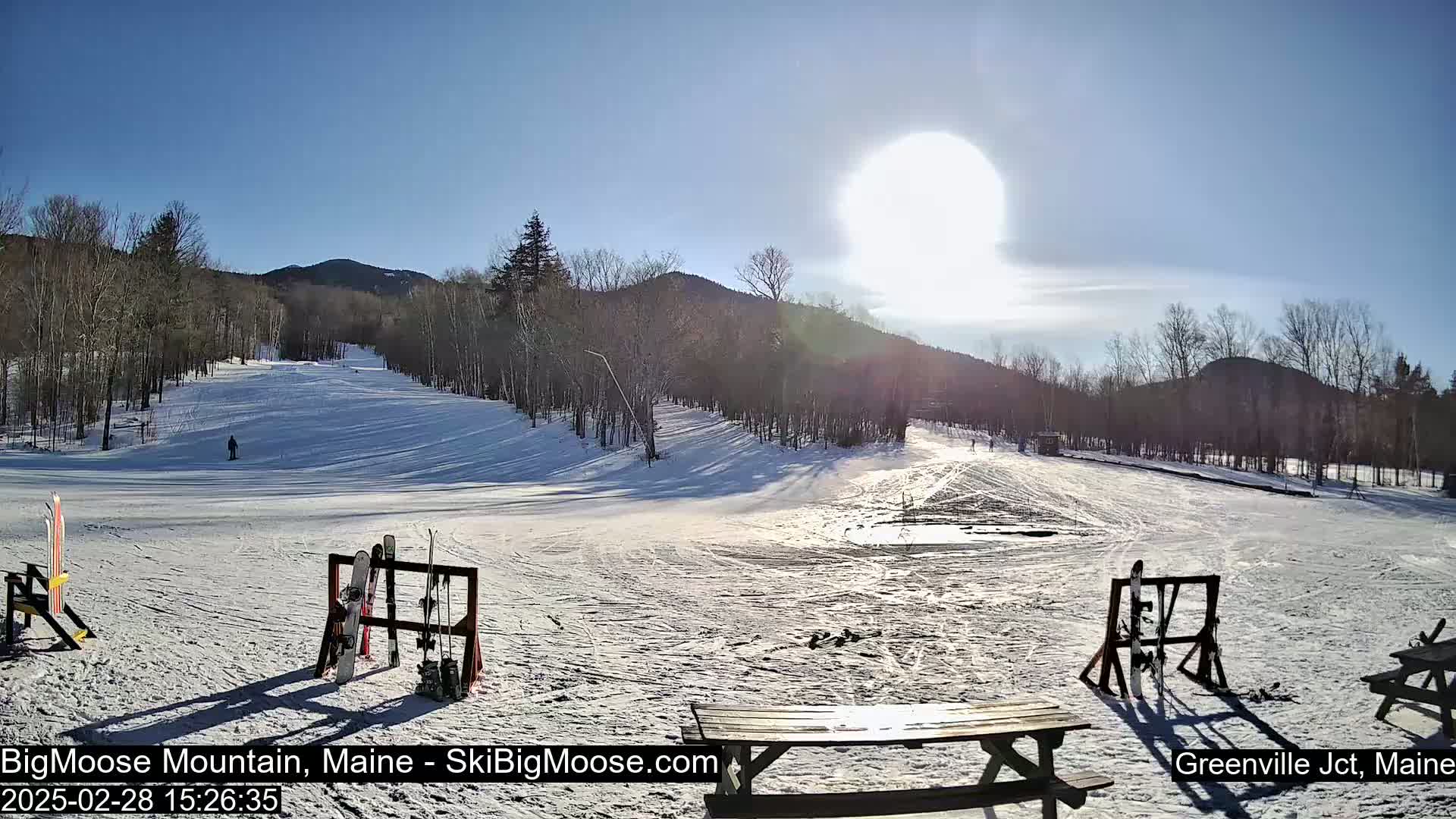 A sunny winter day at a ski slope shows a snow-covered hill with trees and a picnic table in the foreground.