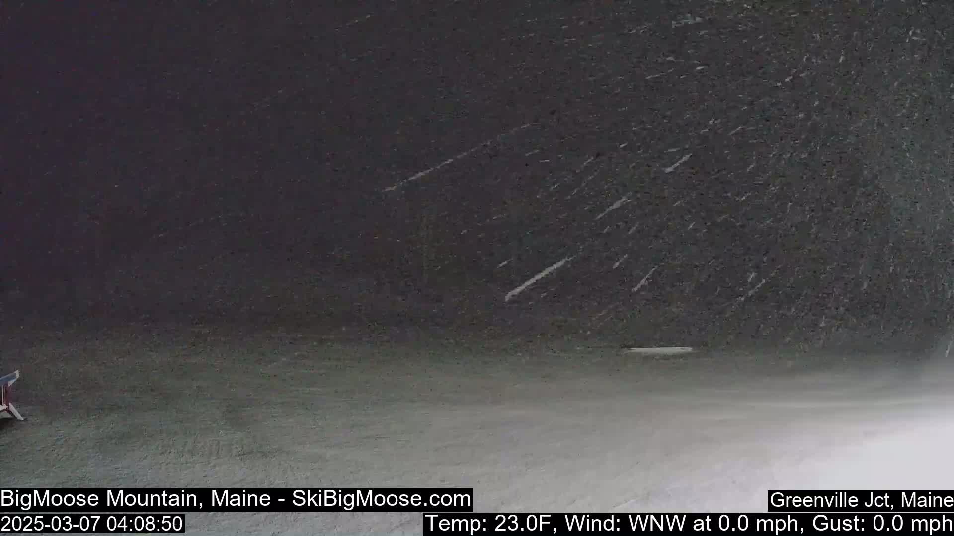 A dark, snowy scene shows light snowfall on a flat, dark-colored ground.