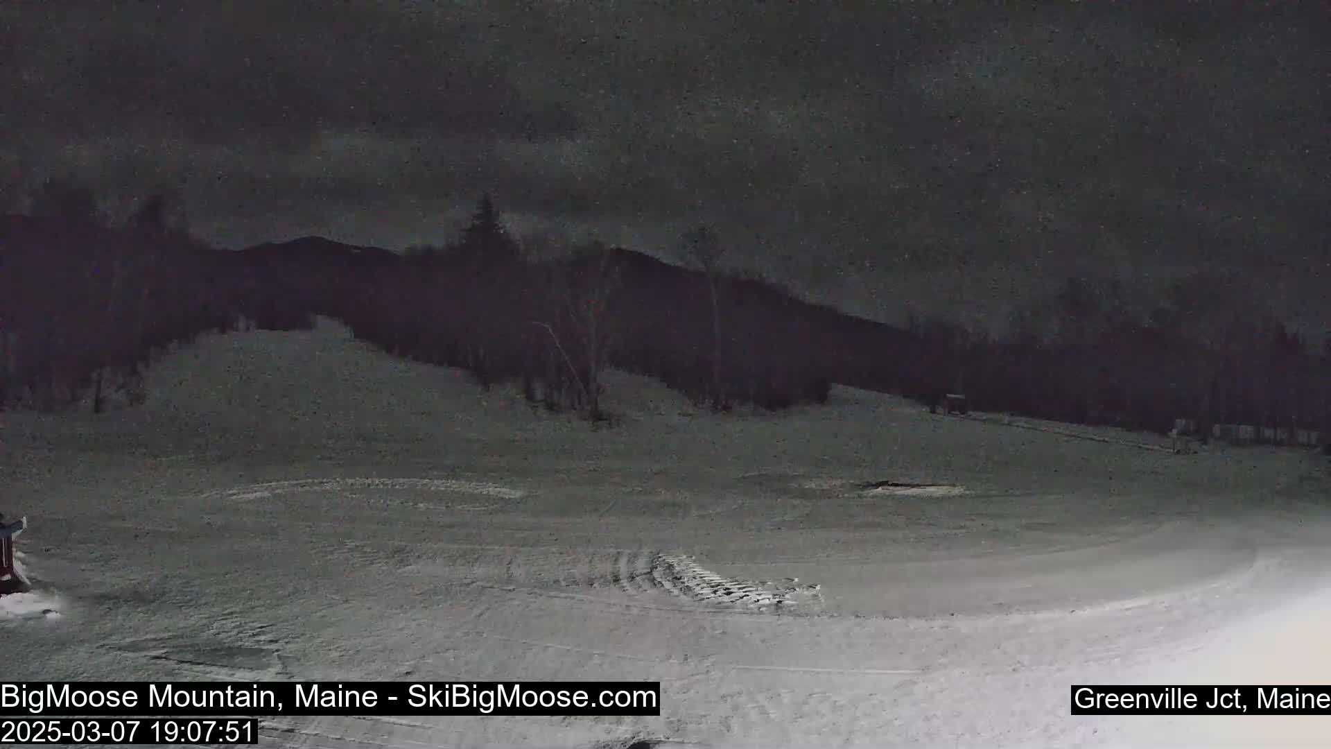 A snow-covered, empty ski slope is visible at night under a dark, cloudy sky.