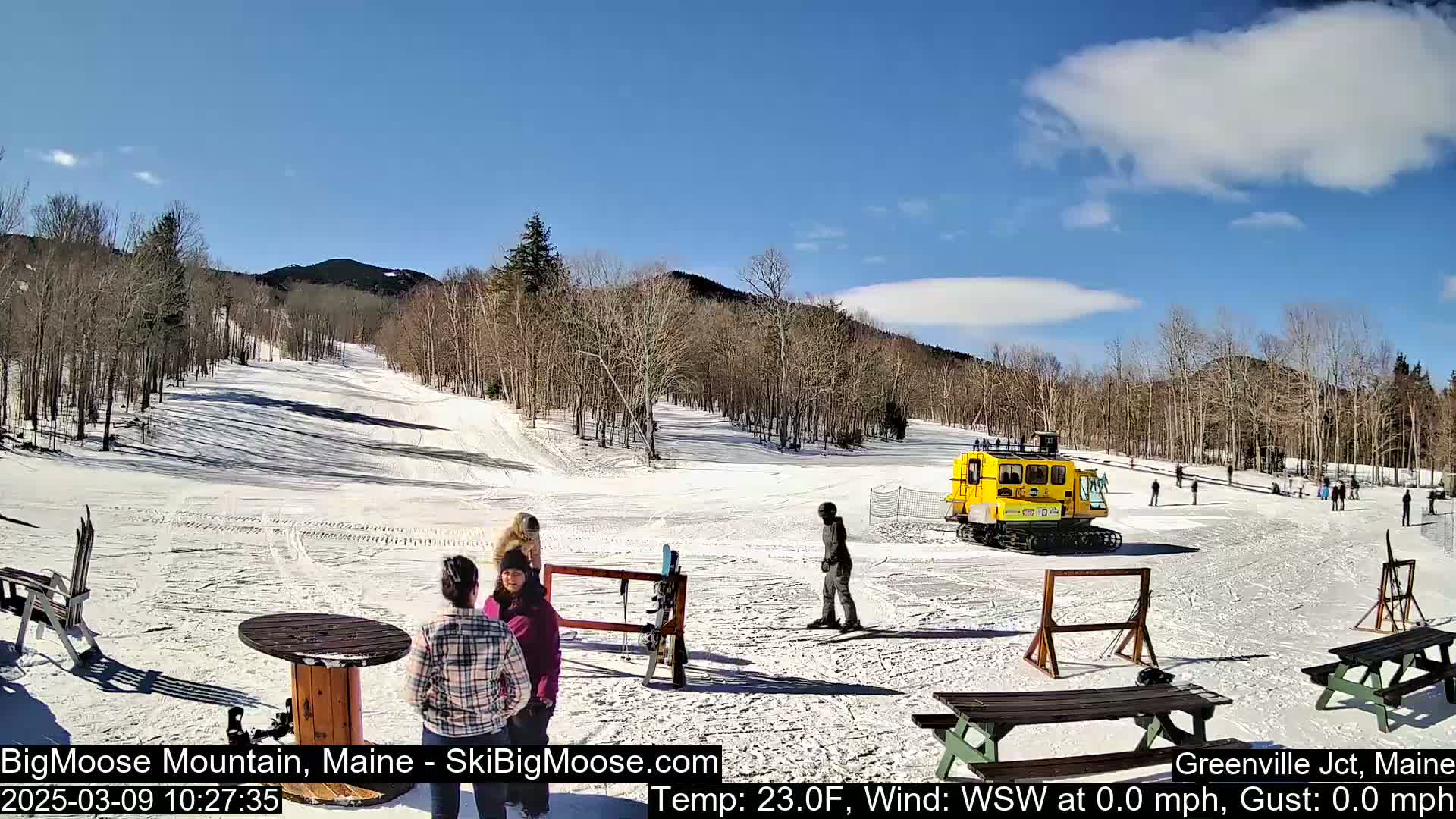 A snow-covered ski slope with several people standing near picnic tables and a yellow snowcat, under a mostly sunny sky.