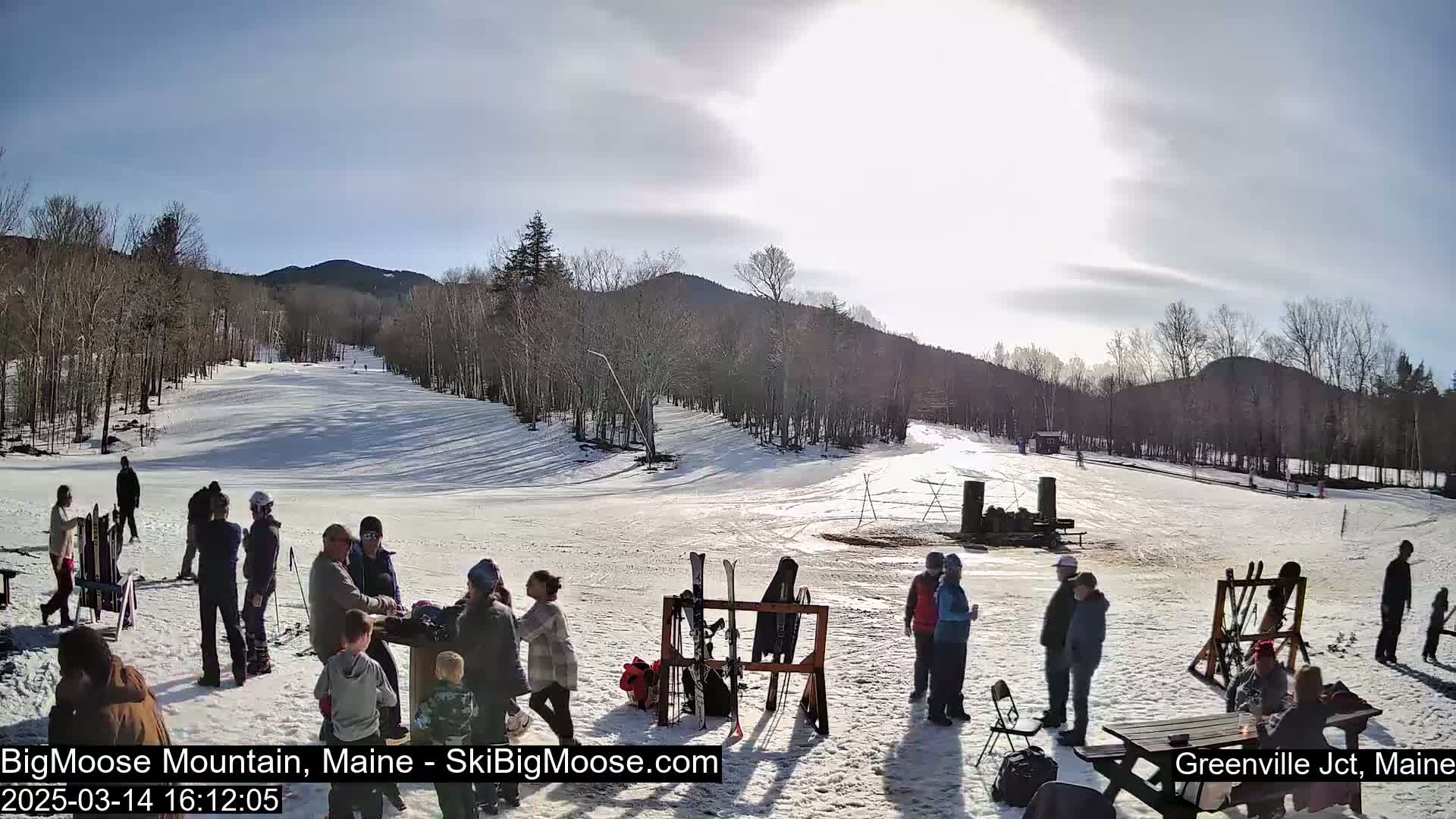 A sunny day at a snow-covered ski resort shows several people gathered near a ski rack and picnic table, with a ski slope and wooded mountains in the background.