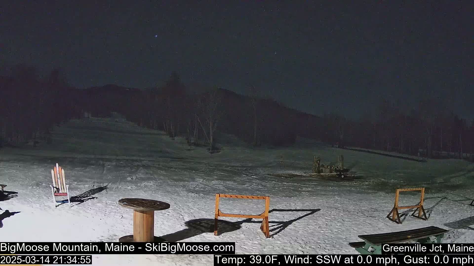 A snow-covered area at night, under a clear, starry sky, features a few pieces of outdoor furniture and some skis leaning against a chair.