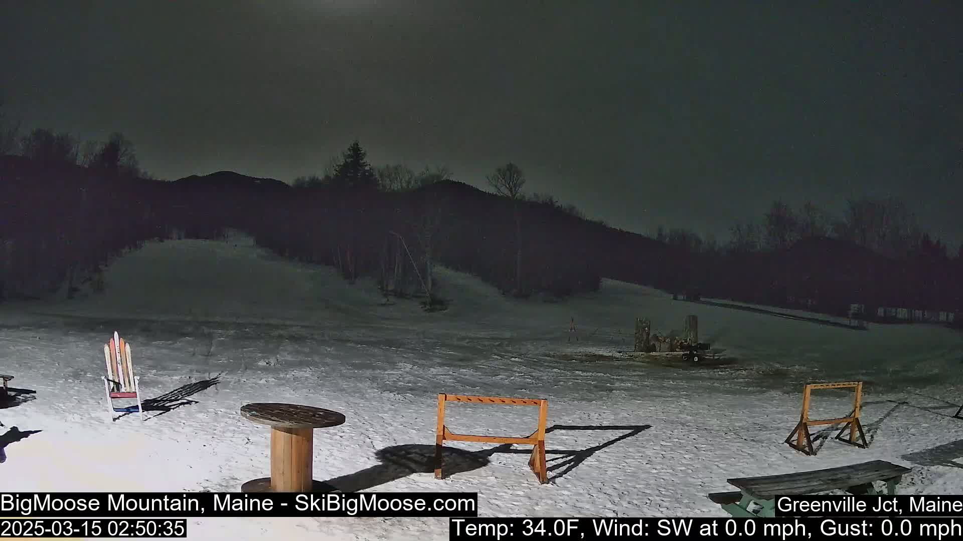 A snow-covered outdoor area at night with a few pieces of outdoor furniture and a distant, dark mountain range under a mostly clear sky.