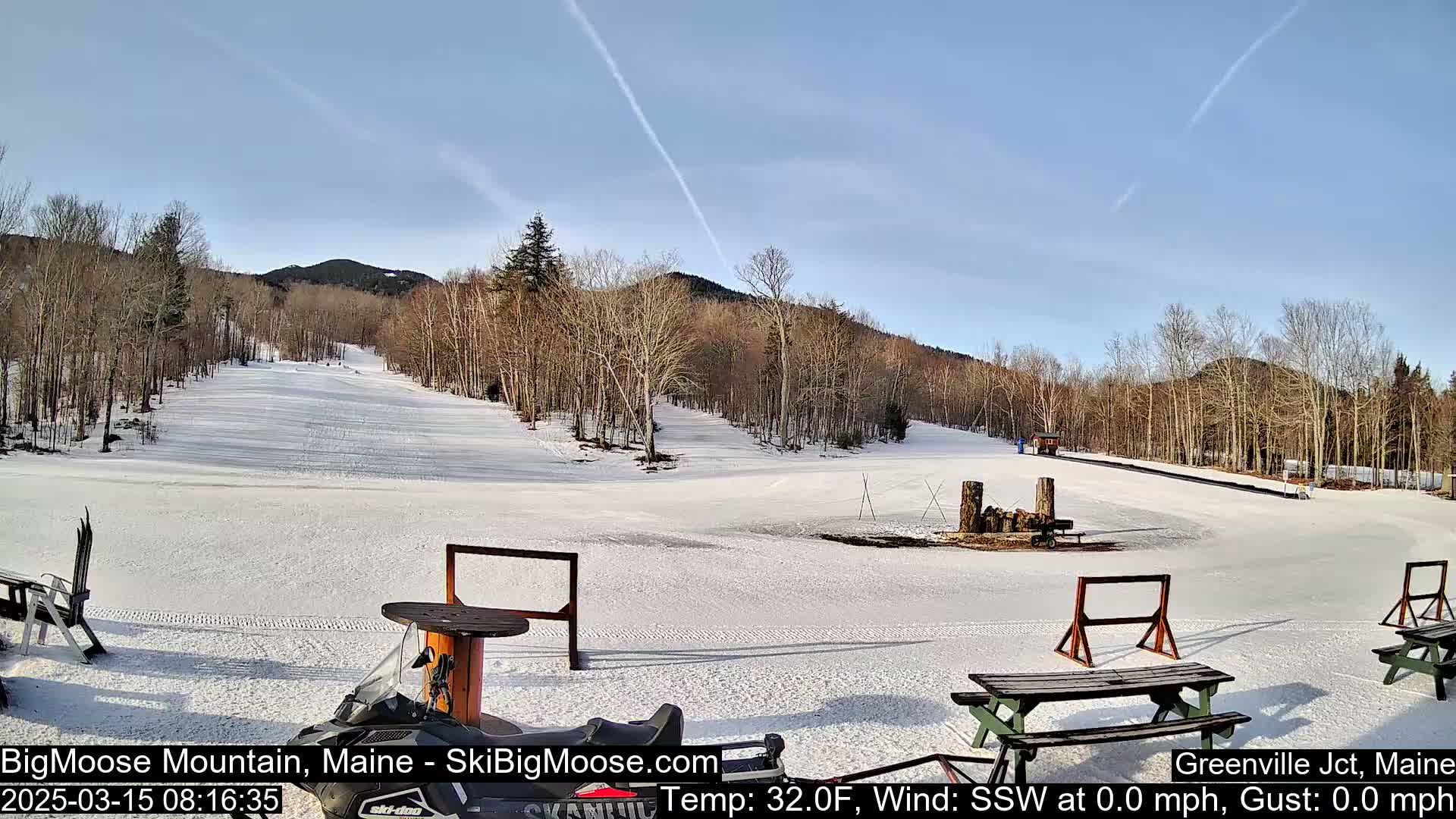 A snow-covered ski slope with sparsely wooded hills in the background under a clear, sunny sky.