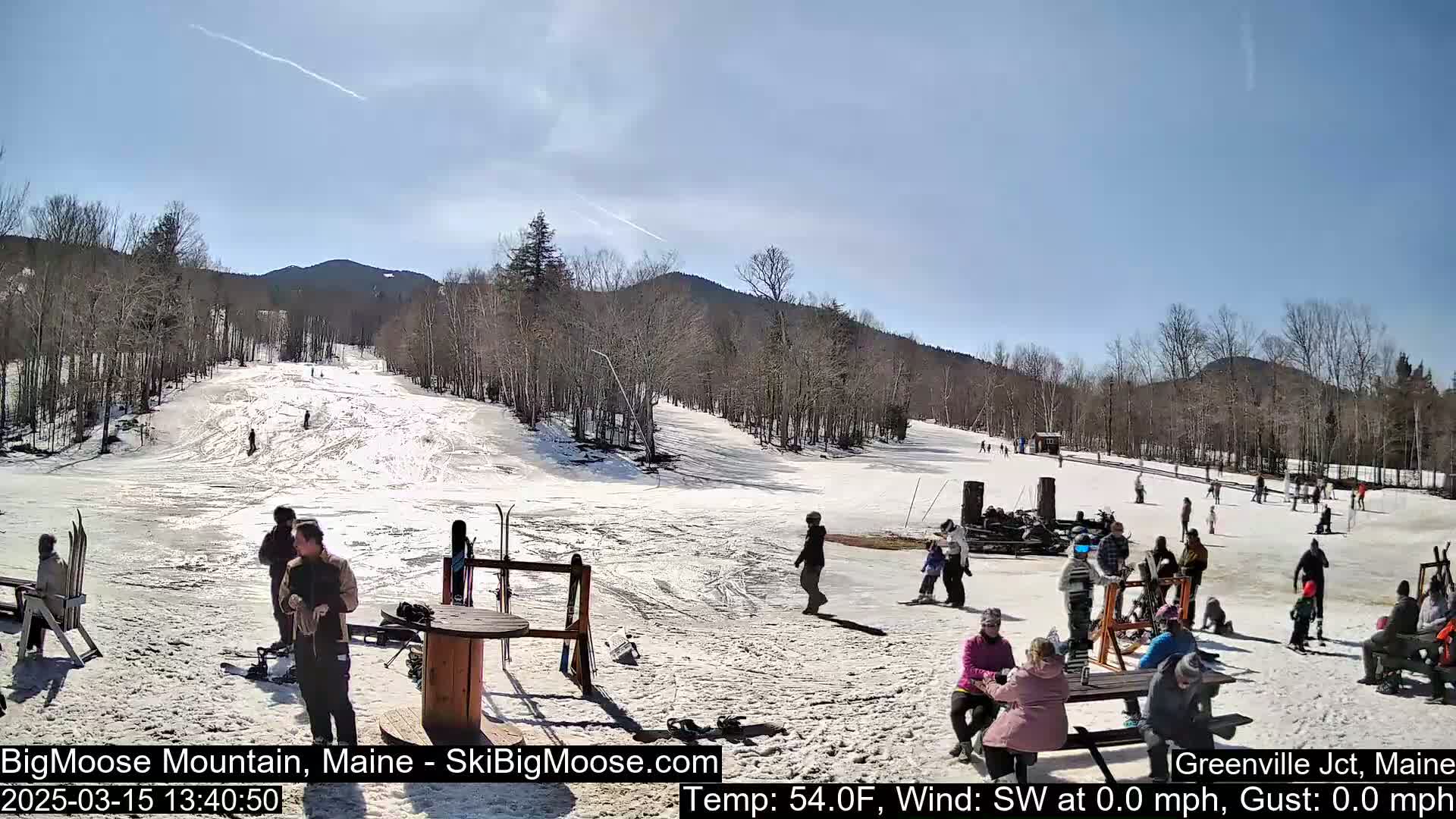 A sunny day at a snow-covered ski resort shows numerous people scattered across the slopes and gathered in a base area.