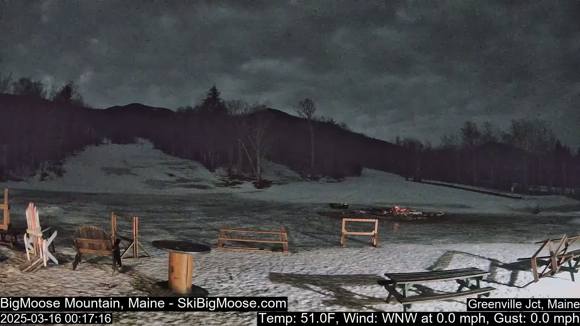 A snow-covered outdoor area with picnic tables and chairs sits under a dark, cloudy night sky, with a hill and trees in the background.