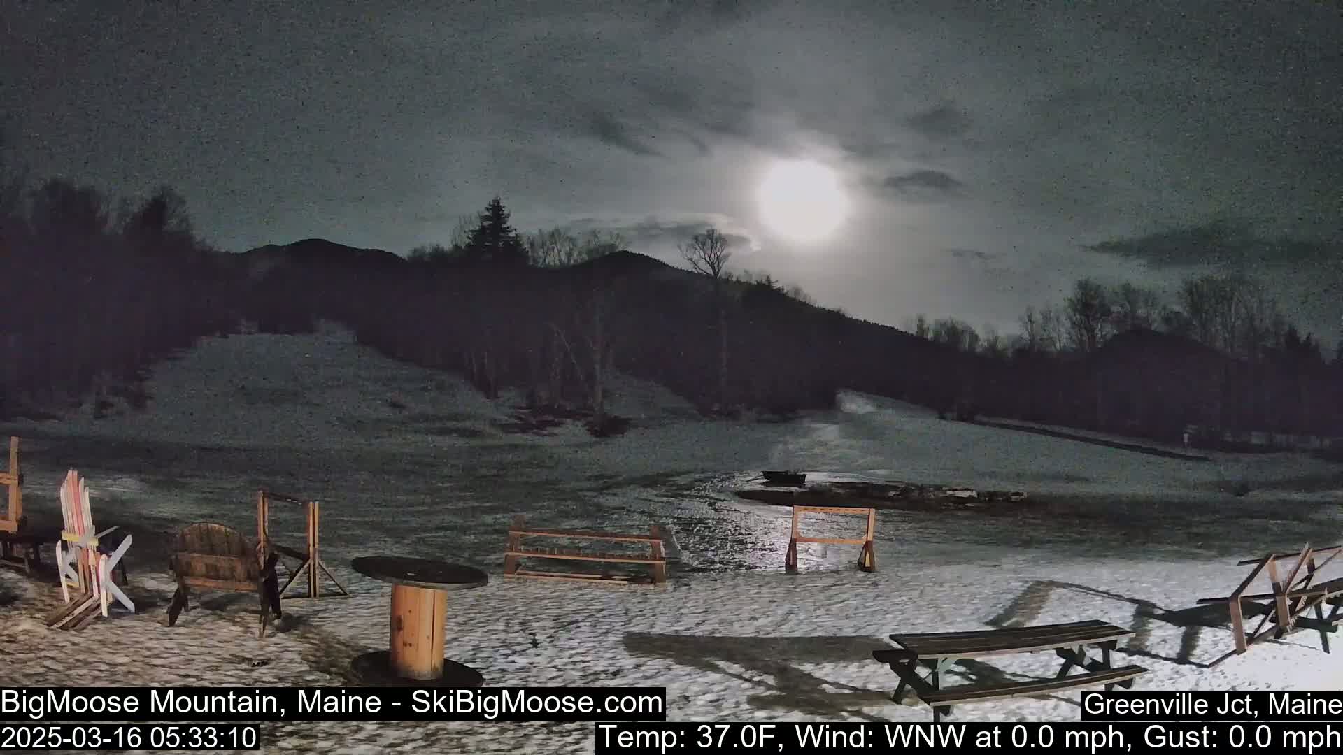 A moonlit snowy scene at night shows a mostly clear sky above a snow-covered area with several park benches and a table, some ski equipment, and a mountain in the background.
