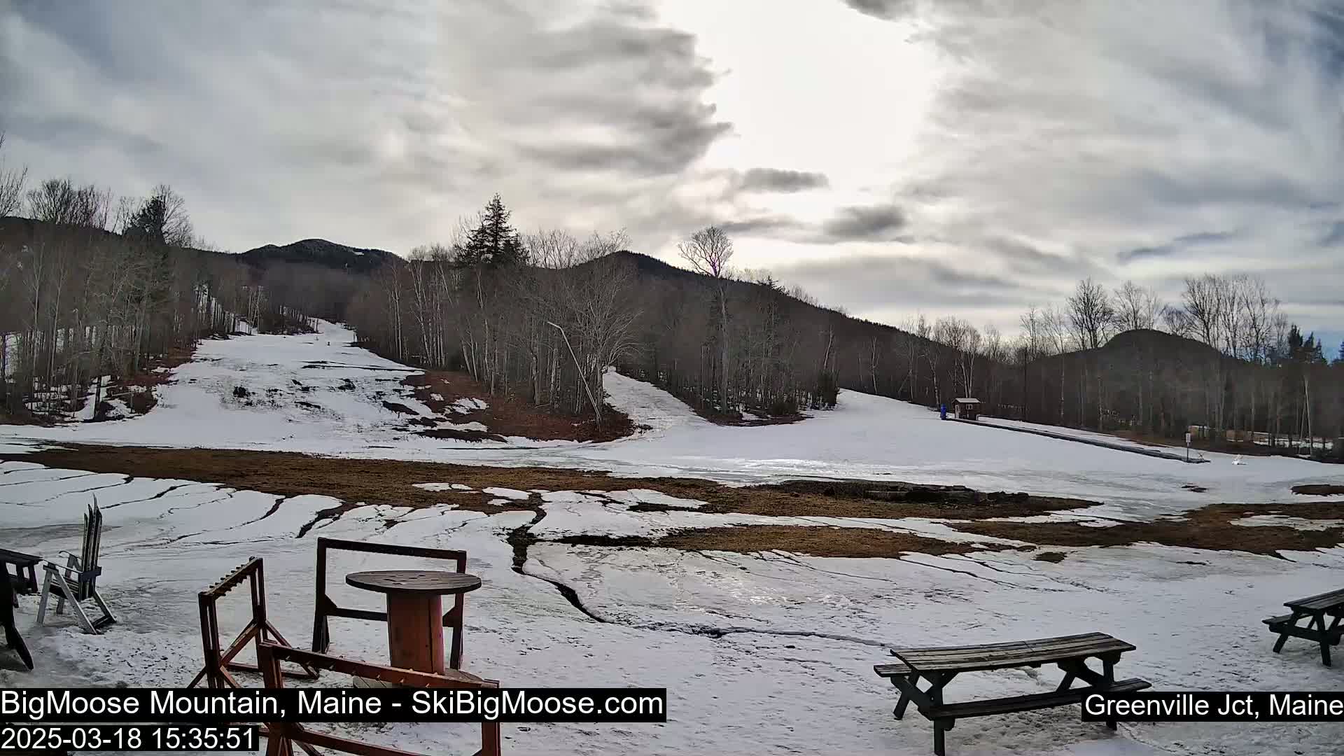 A mostly cloudy day reveals a snow-covered ski slope with patches of brown ground and ice, picnic tables, and bare trees on a hillside.