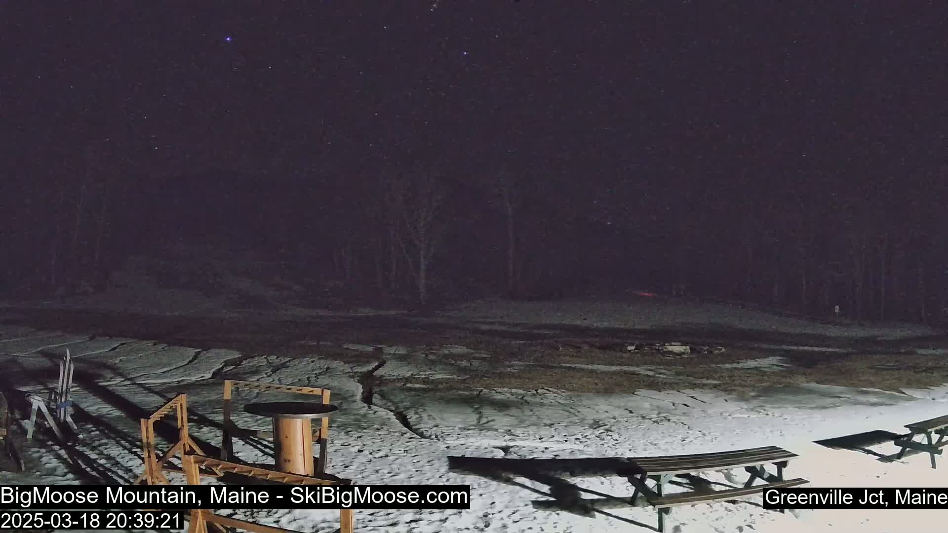 A snow-covered area at night with picnic tables and outdoor furniture under a starry sky.