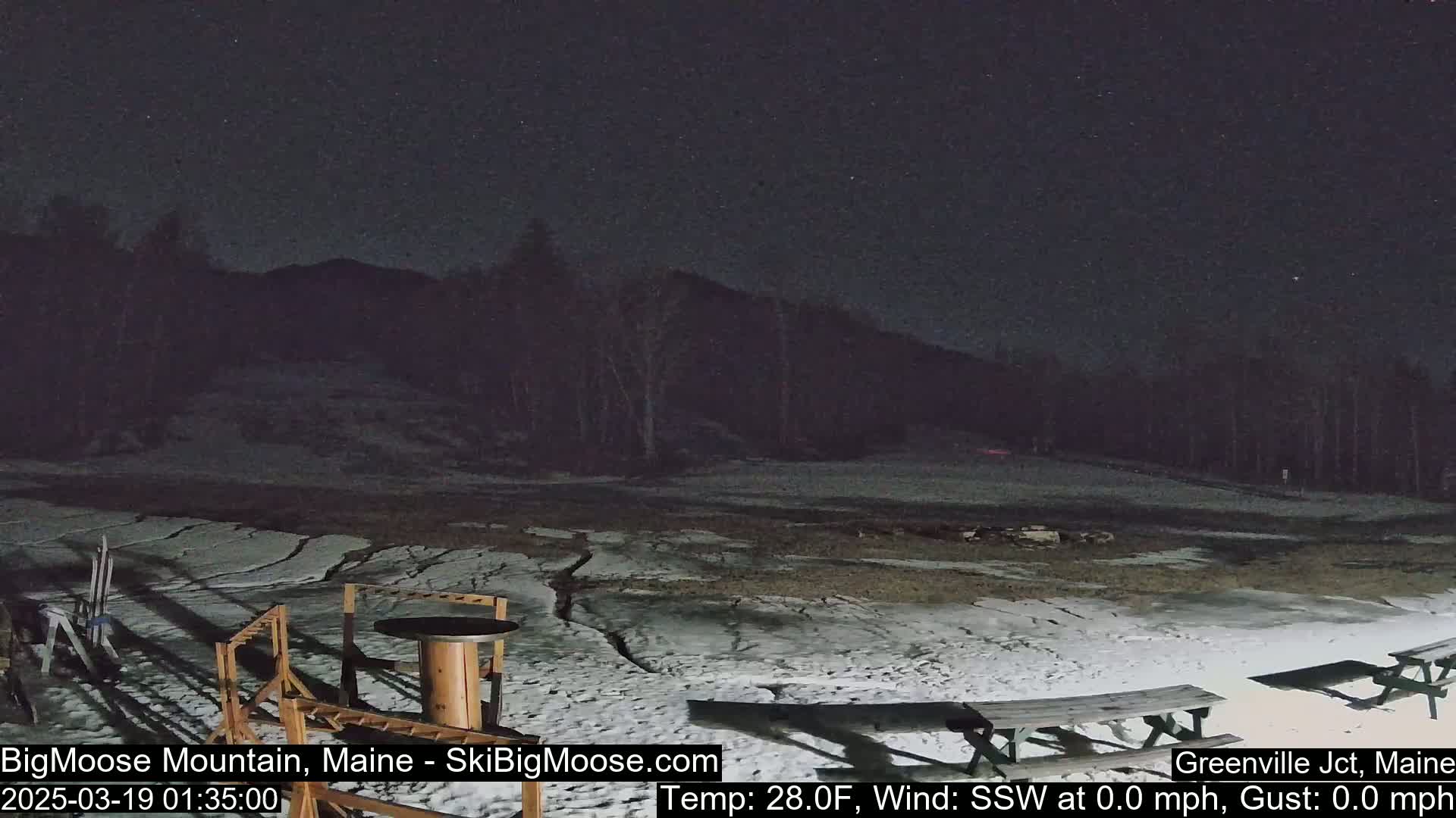 A nighttime view of a snow-covered area with picnic tables and ski equipment, under a clear, starry sky; there is no wind.