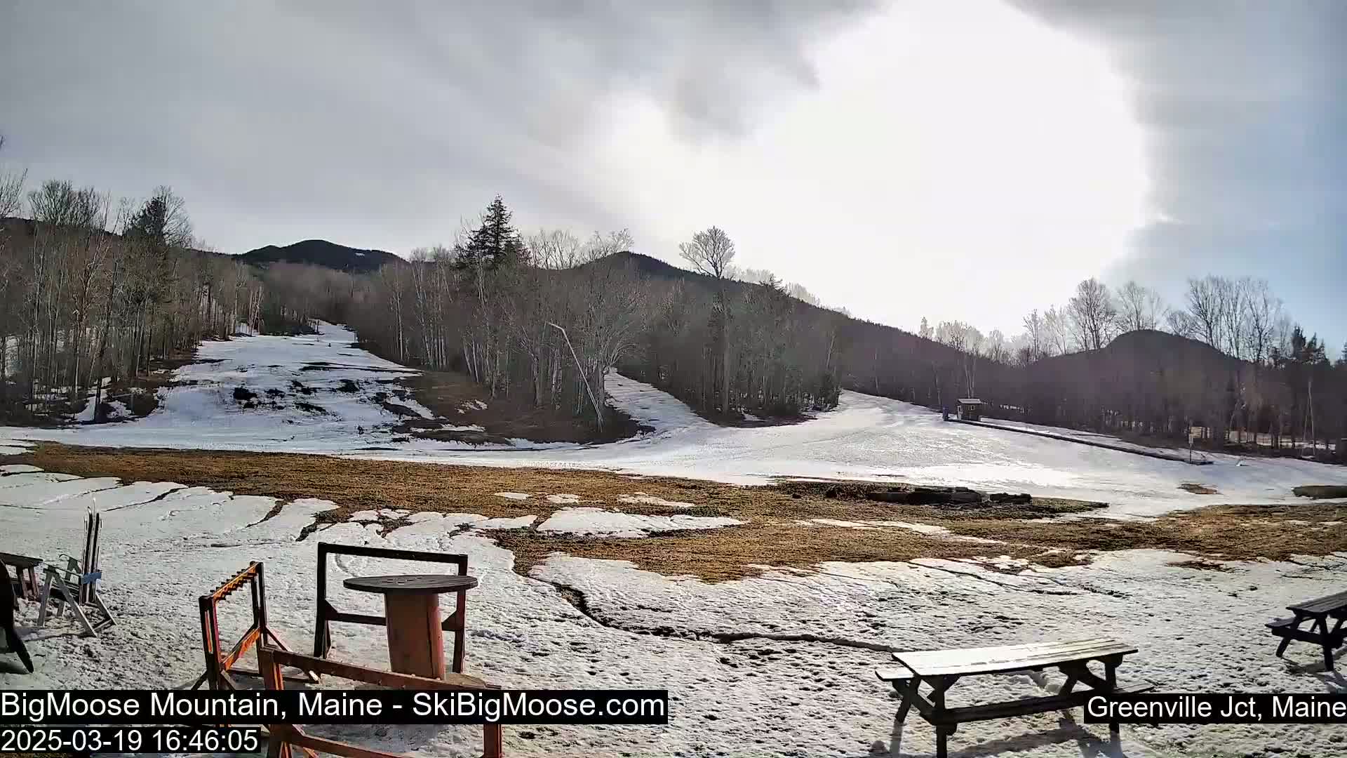 A mostly sunny day shows a snow-covered ski slope with patchy brown ground, surrounded by bare trees and hills.