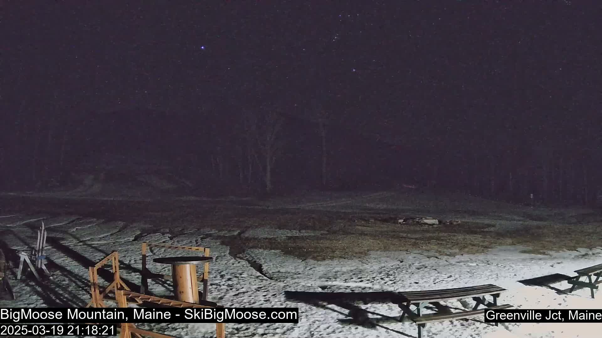 A snow-covered ground at night with a few picnic tables and some equipment under a starry sky.