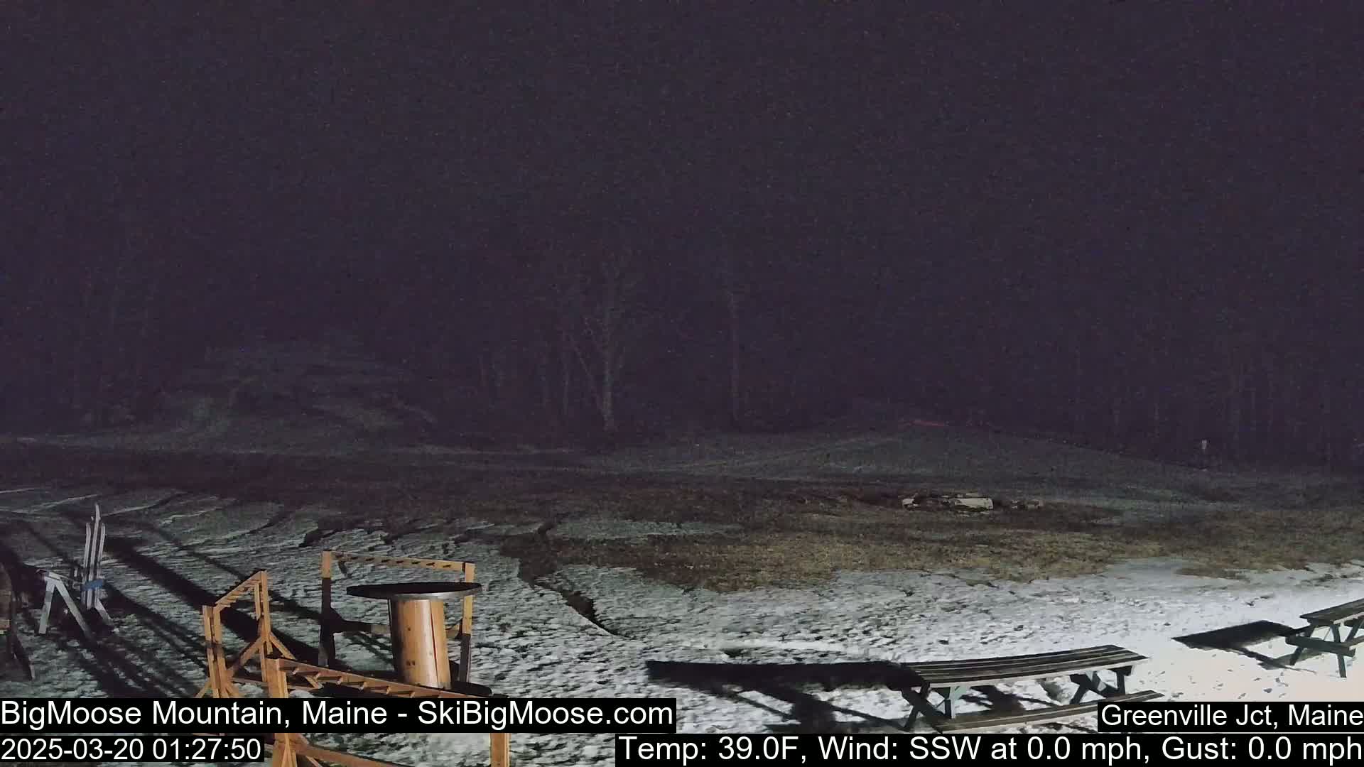 A snow-covered outdoor area at night with picnic tables and some outdoor furniture, under a dark, possibly overcast sky.