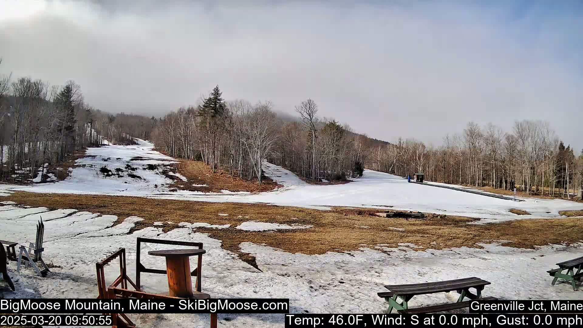 A mostly cloudy day reveals a snow-covered ski slope at the base of a mountain with brown patches of exposed ground and bare, deciduous trees.