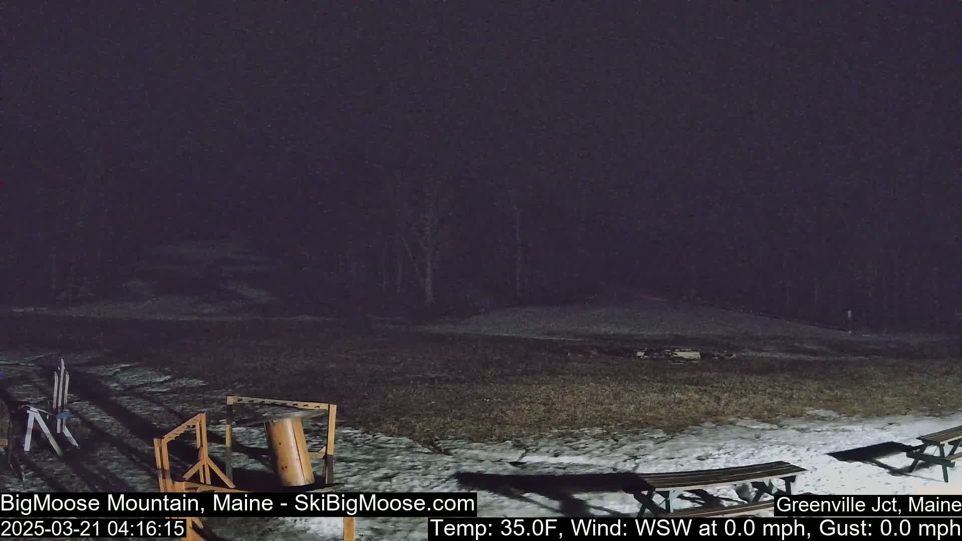 A nighttime view of a snow-covered area with picnic tables and some equipment, under a dark, possibly cloudy sky.