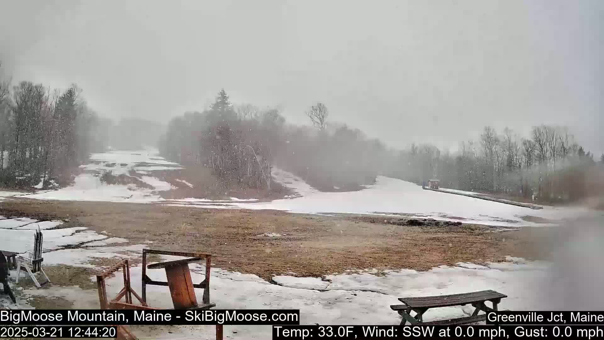 A snow-covered ski slope is visible through falling snow, with bare patches of ground and some wooden picnic tables in the foreground.