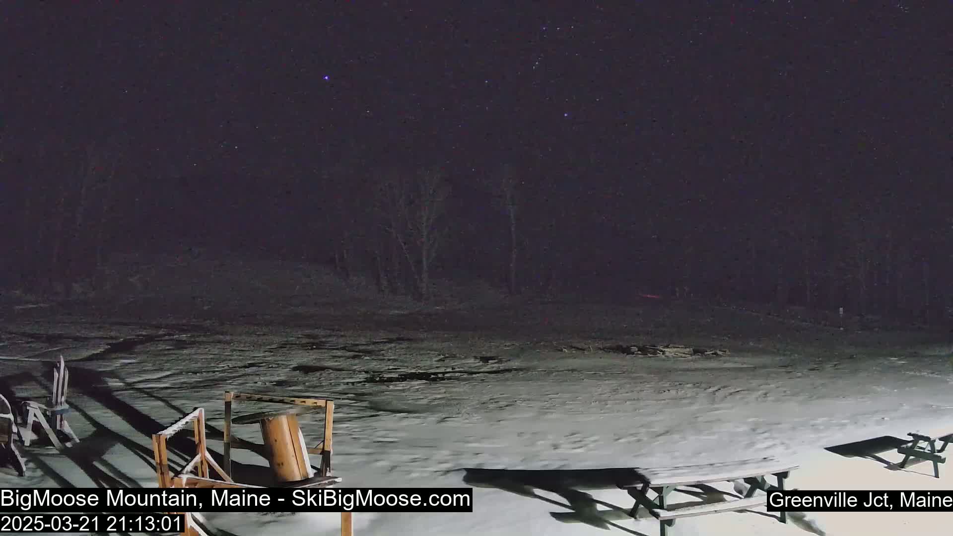A snow-covered area at night with a few bare trees in the background under a starry sky, containing some outdoor picnic tables and other wooden structures.